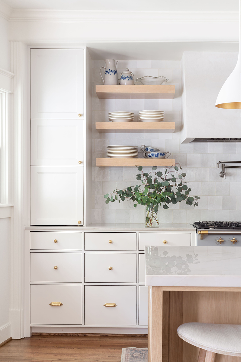 A white kitchen with open wooden shelves holding dishes and jugs, a potted plant, and a white cabinet with gold hardware.