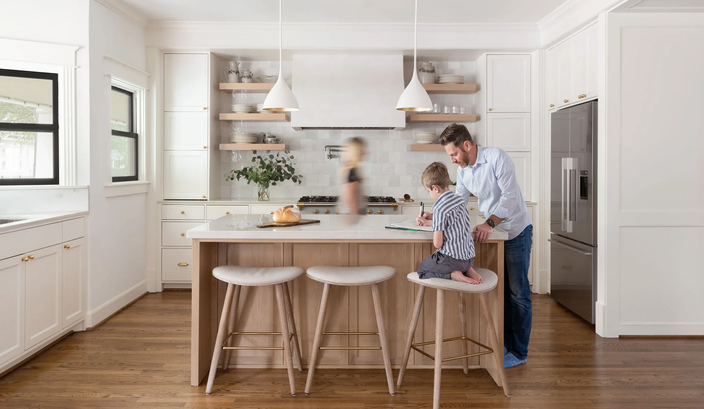 A man and young boy are writing at a kitchen island, with the boy kneeling on a stool. In the background, a blurred child walks past in a bright white kitchen with open shelves, a potted plant, and a gas range.