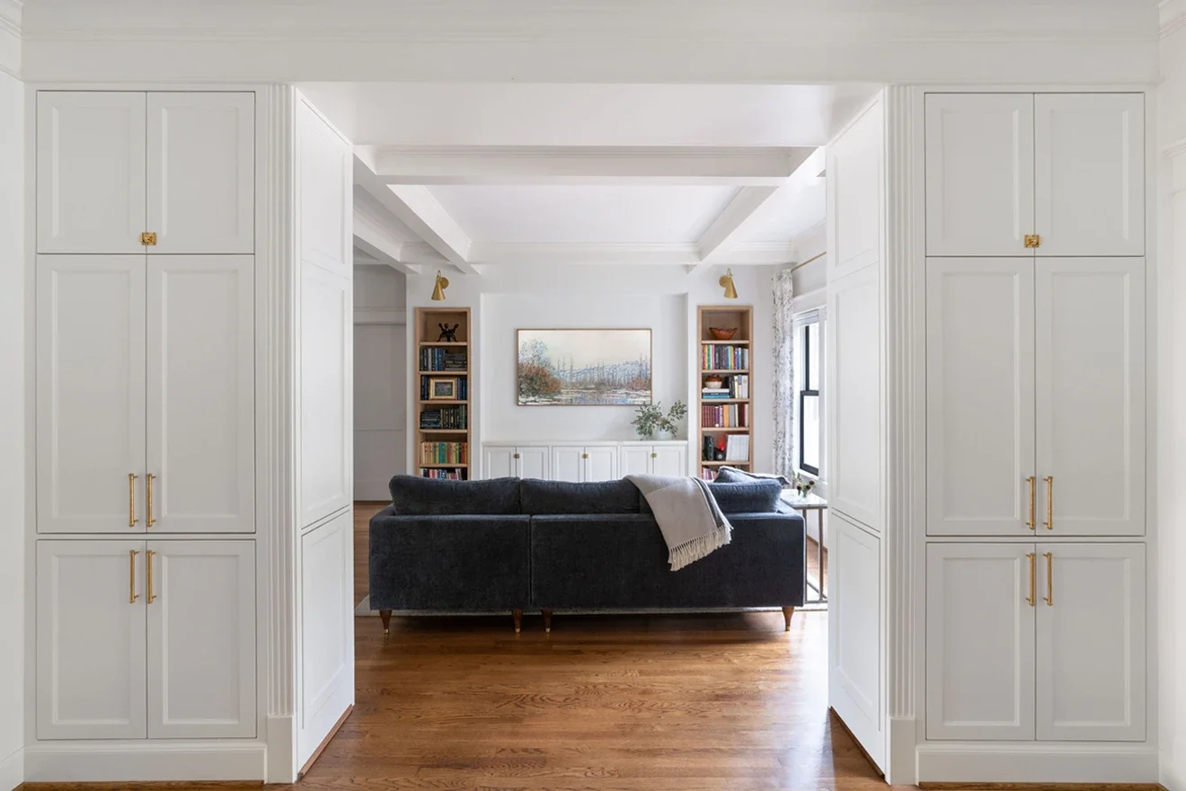 Living room with white walls, gold accents, and wooden floors, featuring a black sofa, bookshelves, a framed landscape painting, and large windows.