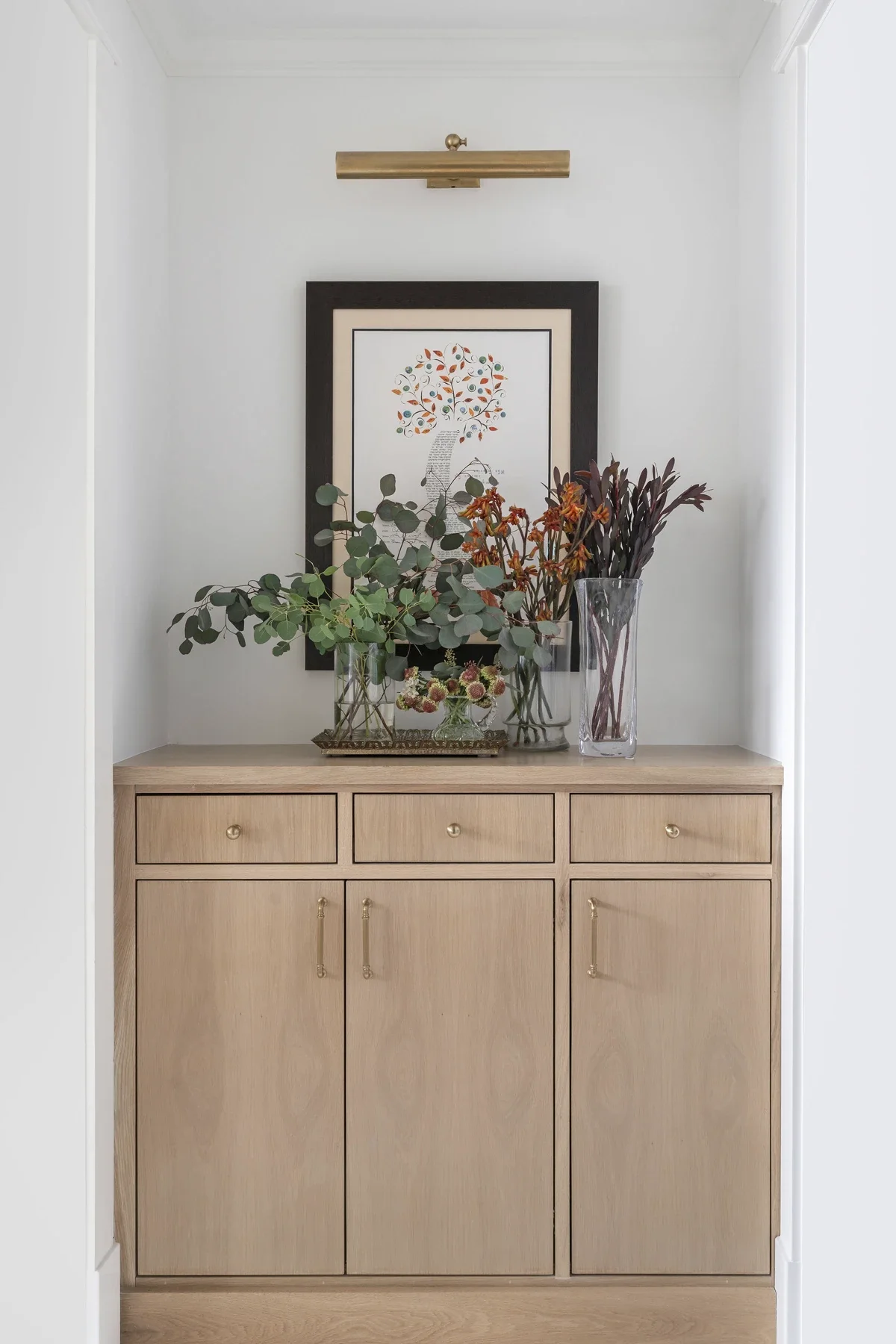 A light wood sideboard with three drawers and two cabinet doors, decorated with a tray of various green and reddish plants, and a framed artwork of a tree with colorful leaves on the wall behind, in a white-walled room.