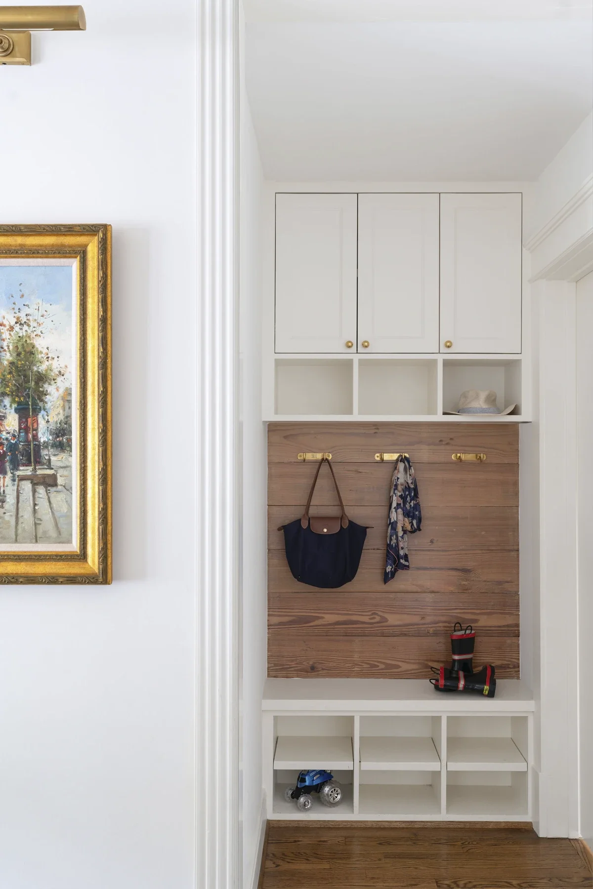 Entryway with white cabinets, wood panel, hooks with bags, a hat, and small cubbies with a toy truck.