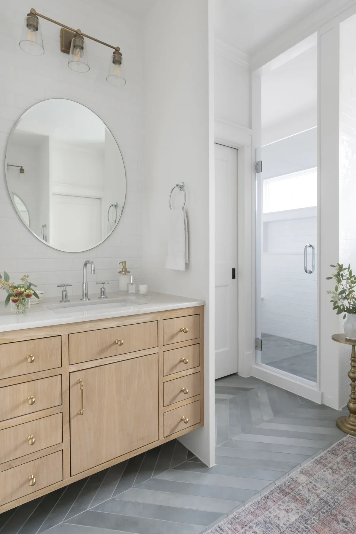A bright bathroom with a wooden vanity, round mirror, wall-mounted light fixture, and a glass-enclosed shower.