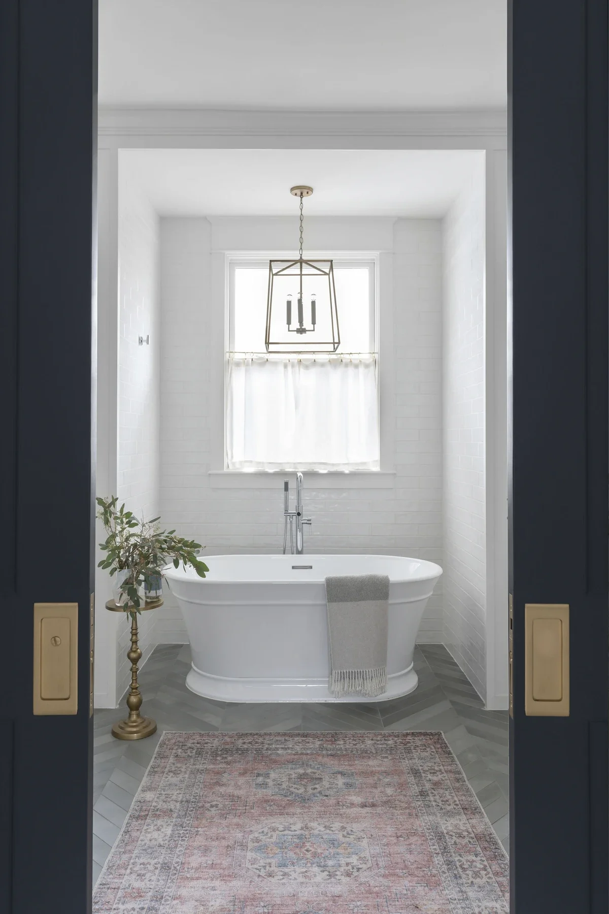 Bathroom with a bathtub in front of a window, a hanging light fixture above, and a plant on a stand to the left.