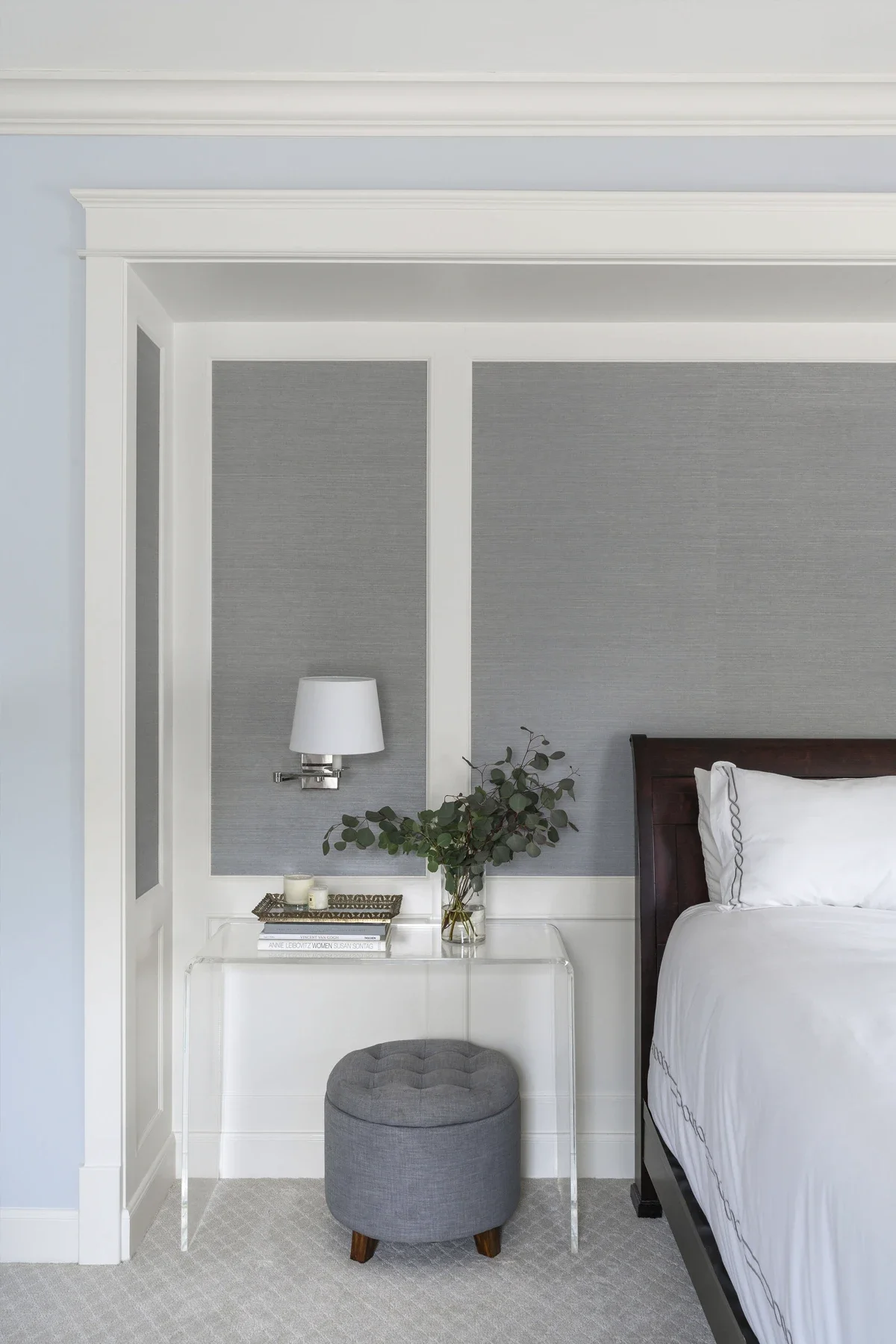 A bedroom with a white bedside table, a lamp, a small potted plant, hardcover books, a gray tufted ottoman with wooden legs, and a bed with white bedding and embroidered pillows. The wall behind has gray textured panels with white trim.