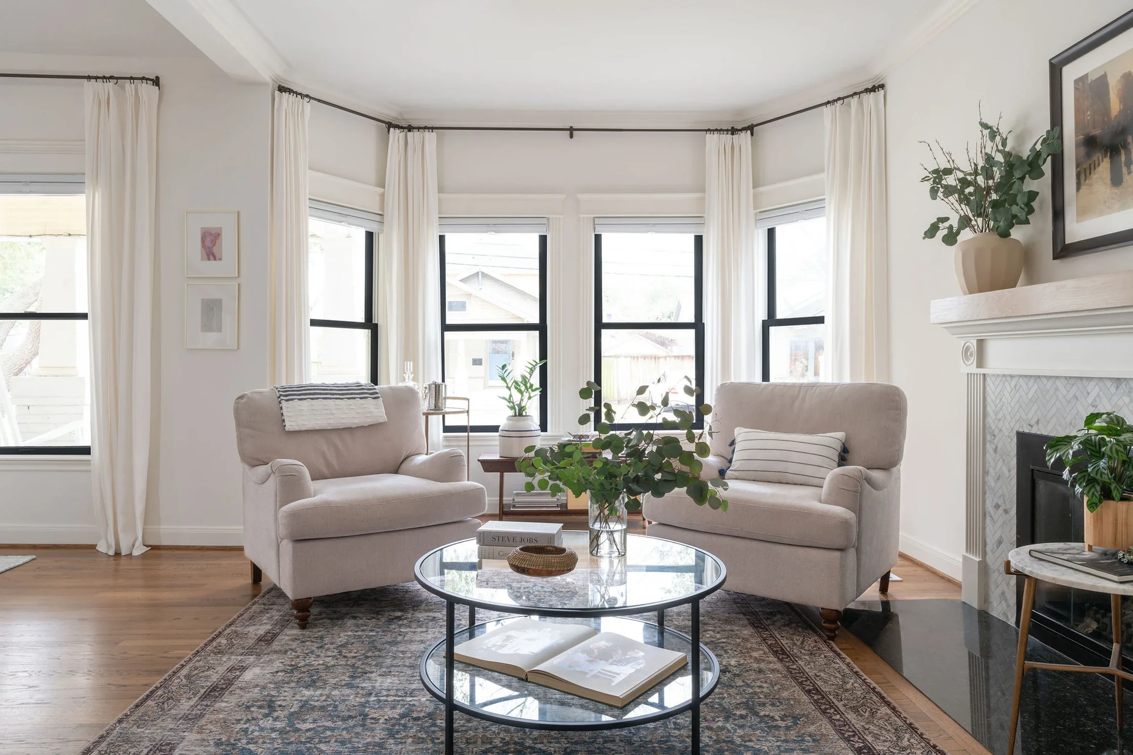 Bright living room with large bay windows, white curtains, beige armchairs, glass top coffee table, and decorative plants.