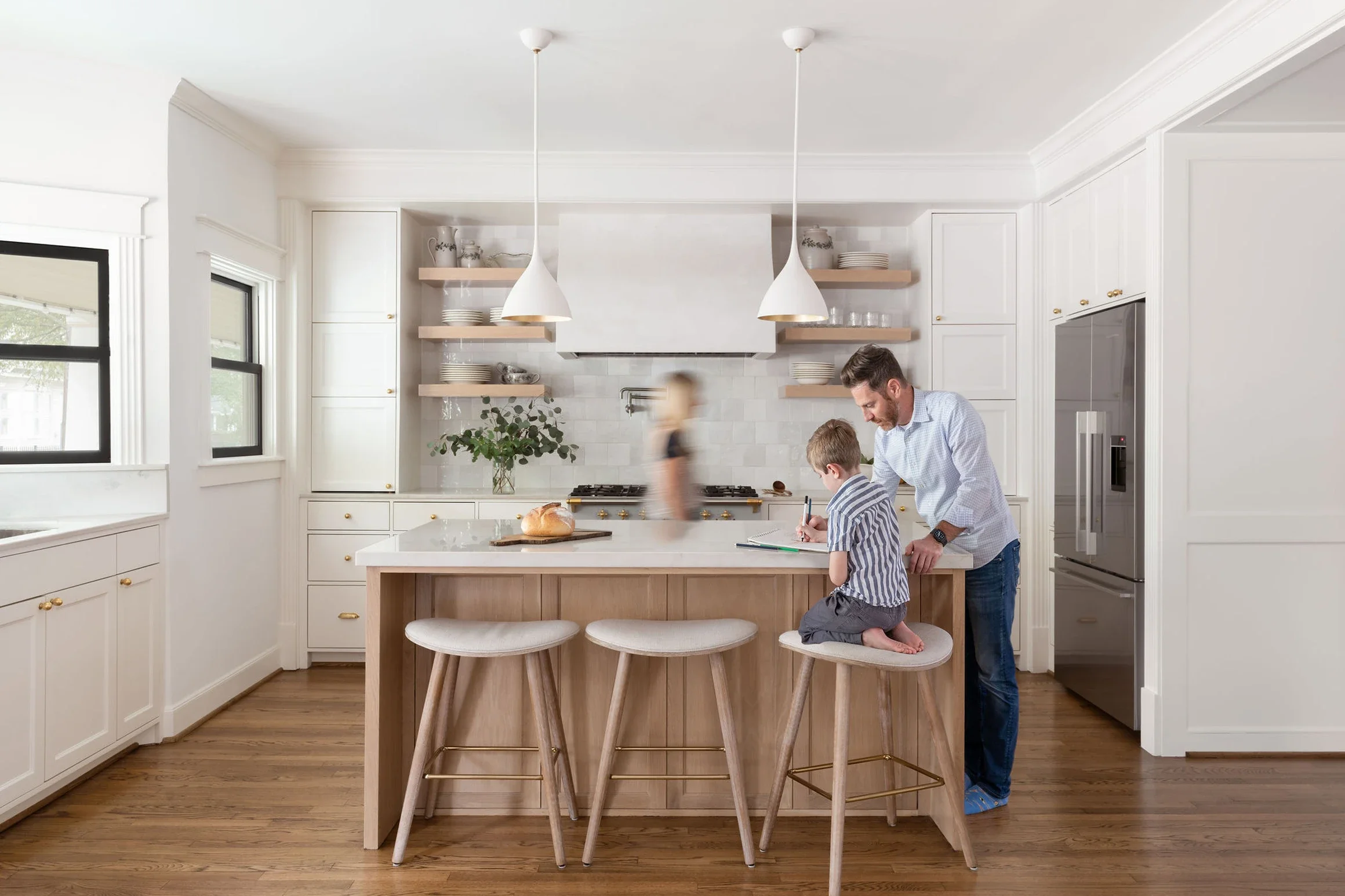 A father and son sitting on kitchen stools at a kitchen island, with a woman walking by in the background of a bright, modern white kitchen.