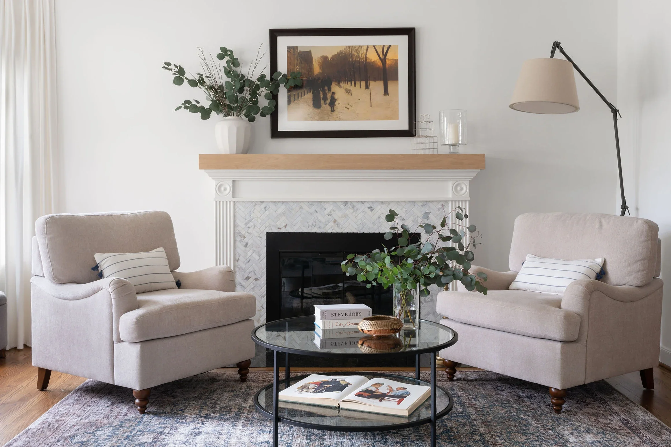 Living room with two beige armchairs, a black glass coffee table, a white fireplace with decorative molding, artwork above the fireplace, a large vase with eucalyptus branches, and a tall floor lamp.