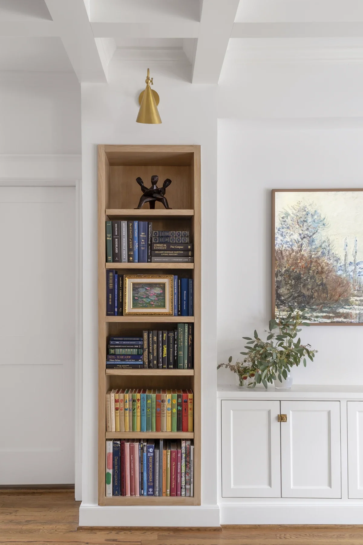 A white wall with a natural wood bookshelf filled with books and a framed painting, a brass wall light, and a white cabinet with a potted plant next to it.