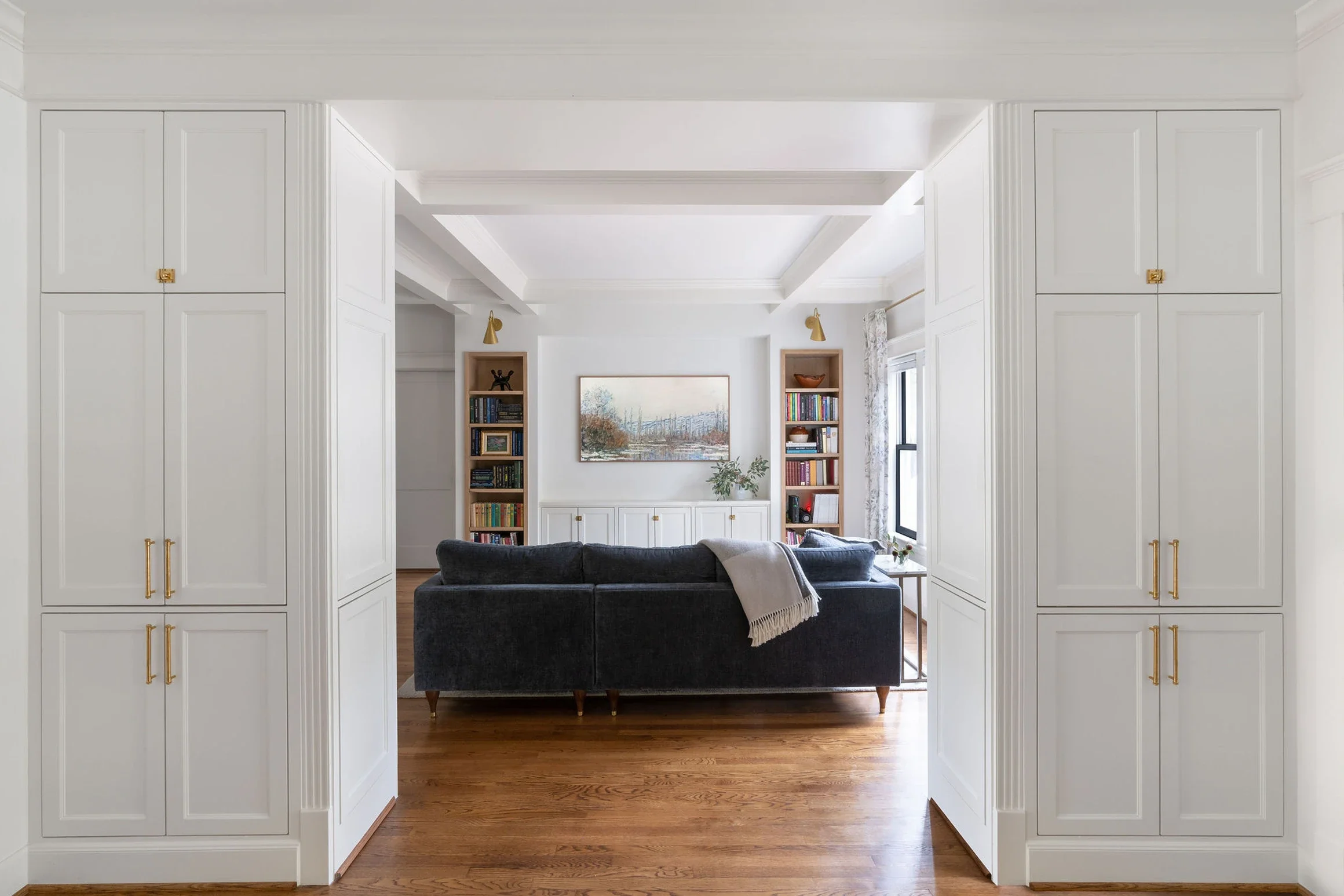 Living room with white built-in cabinets, hardwood floors, a dark gray sofa with a blanket, and bookshelves on either side of a large window.