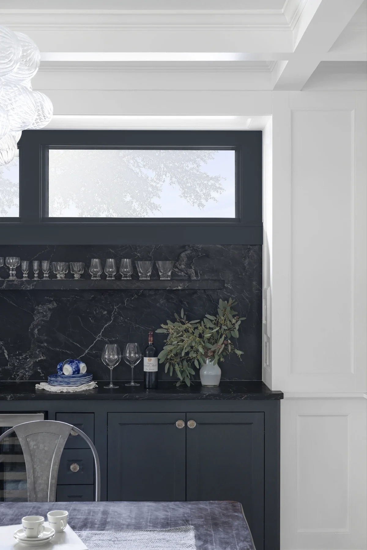 A corner of a dining room with a dark blue cabinet, marble backsplash, and a window. The cabinet holds wine glasses, a bottle of wine, a vase with greenery, and a set of small dishes. Part of a dining table with cups and a table cloth are visible in 