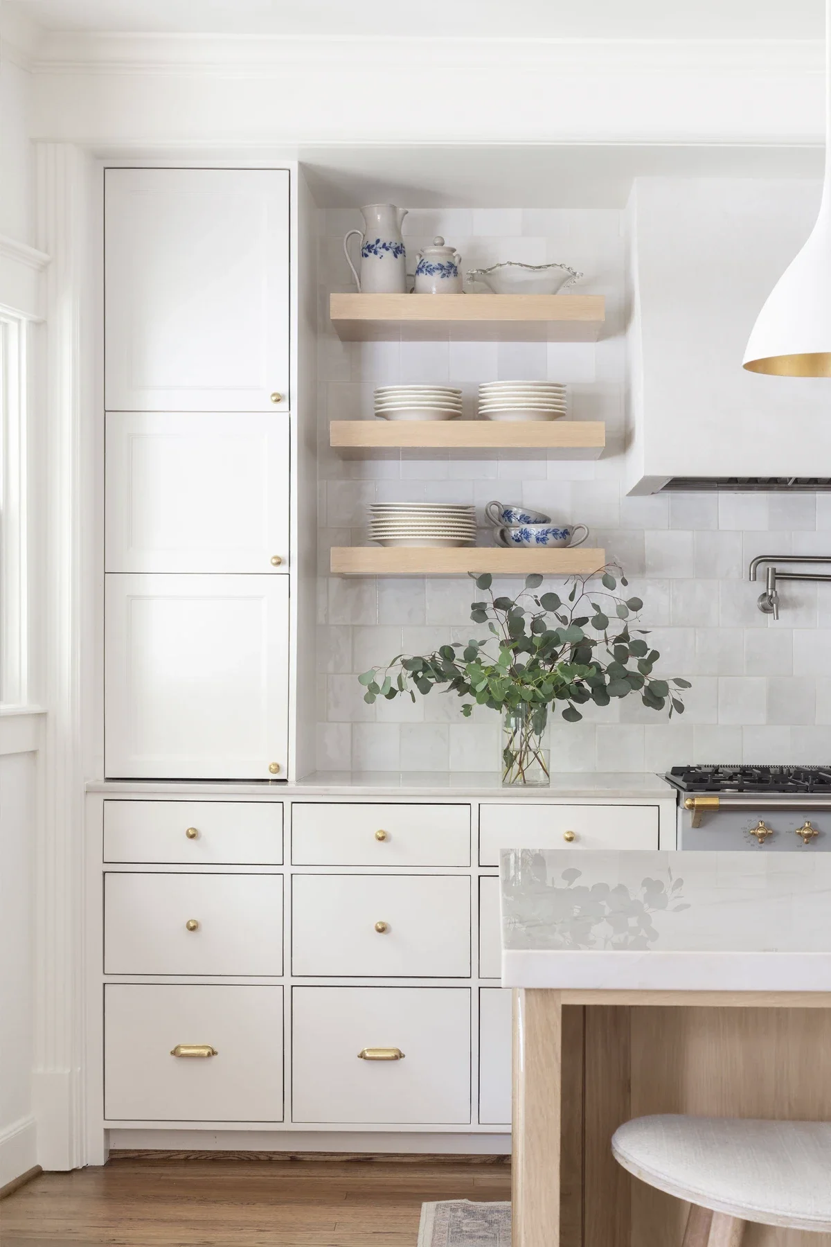 White kitchen with open wooden shelves holding plates, bowls, and teapots, a vase with greenery, and a built-in cabinet with gold knobs.