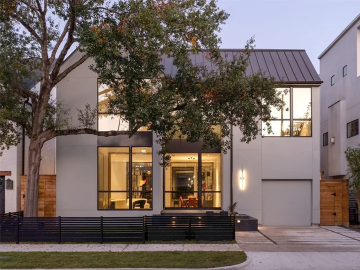Modern two-story house with large glass windows and a metal roof, front yard with a tree and black metal fence, during dusk.