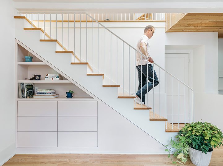 A man walking down a staircase in a modern home with white walls and wooden accents, next to a built-in bookshelf and a large potted plant.