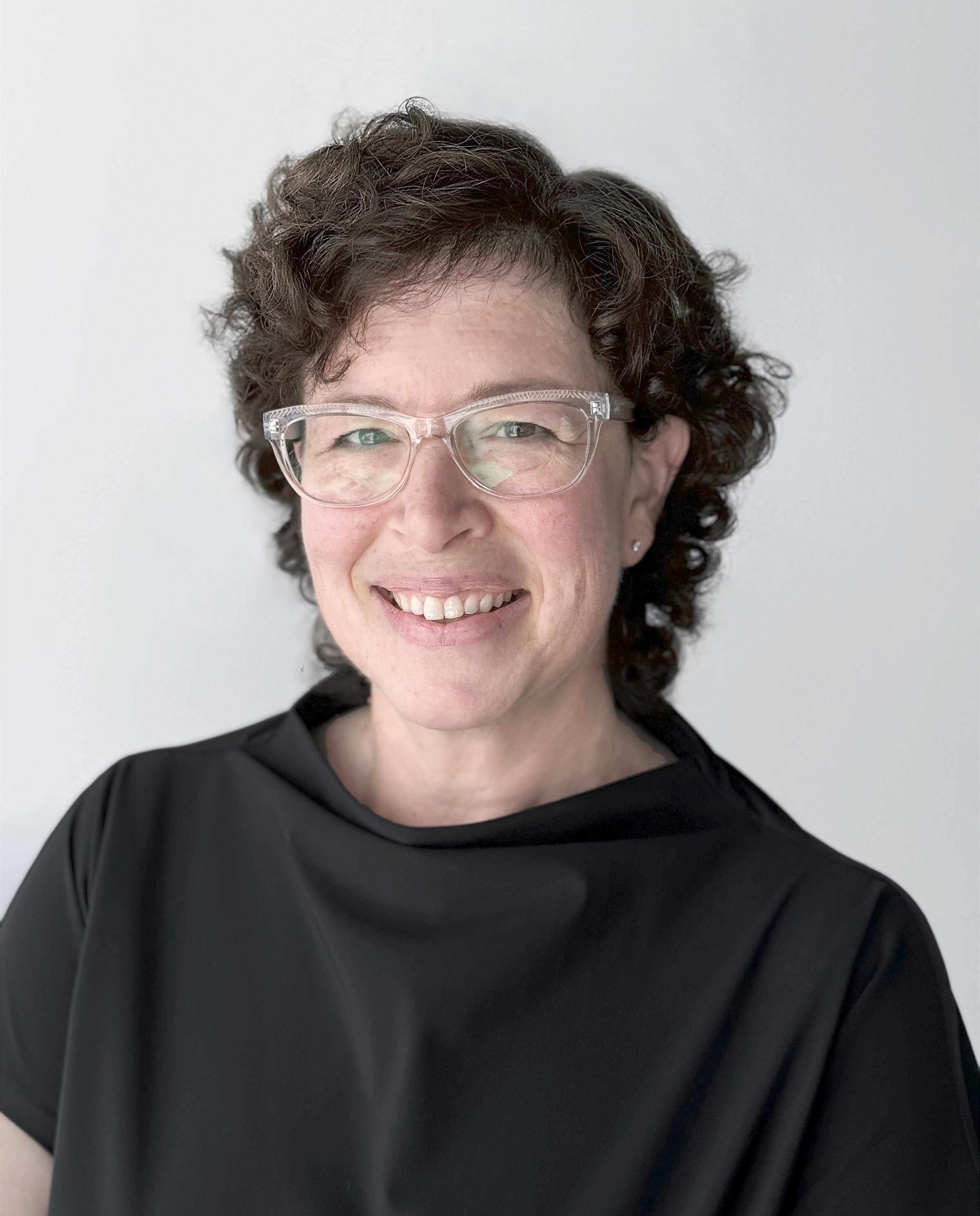 A woman with curly dark hair and glasses smiling in front of a plain light background.