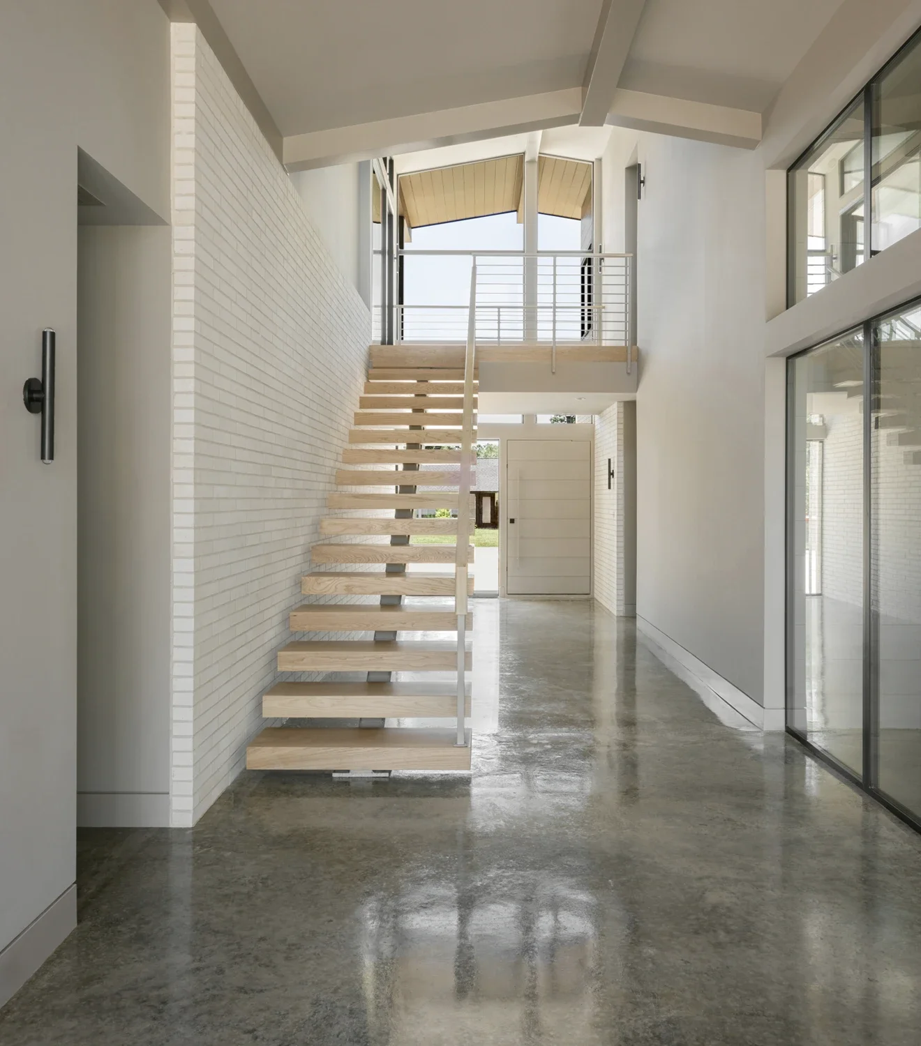 Interior view of a modern, minimalist house with concrete floors, white walls, a wooden staircase, large windows, and a balcony at the top of the stairs.