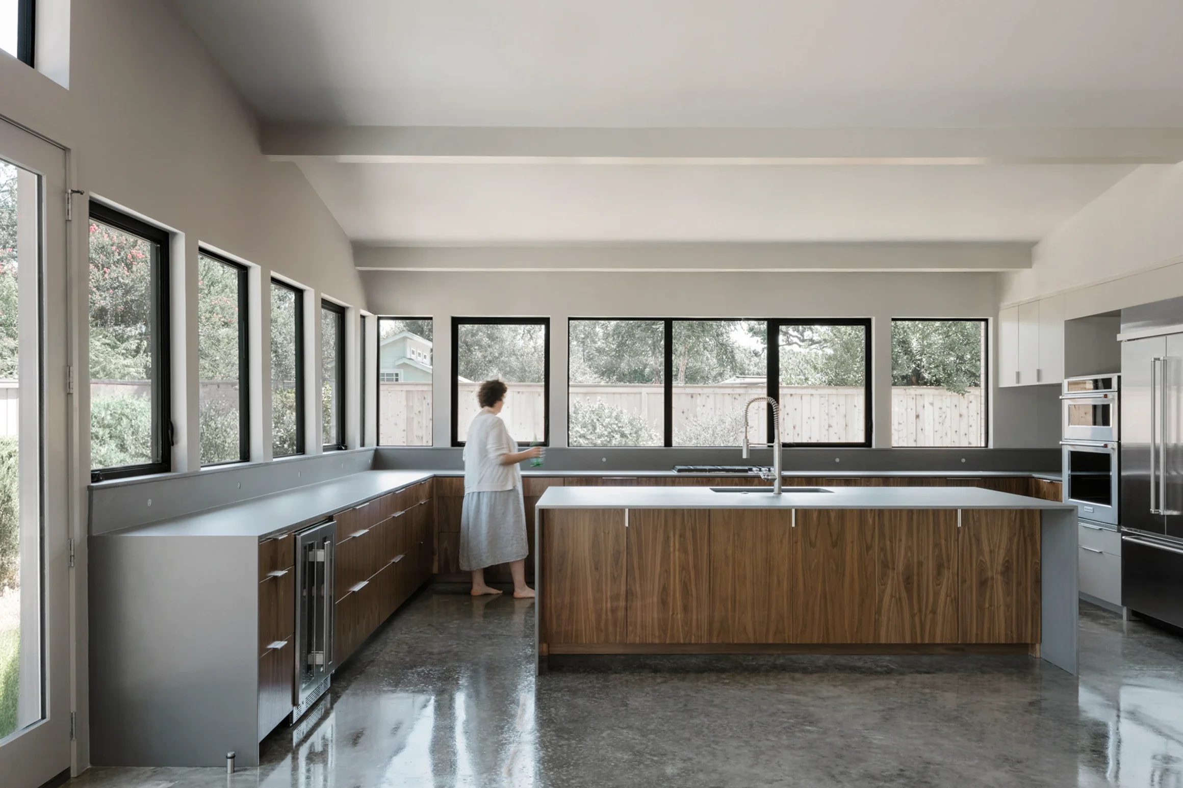 Modern kitchen with wooden cabinets and a large island, woman standing by window with a glass in hand, multiple windows letting in natural light, polished concrete floor.