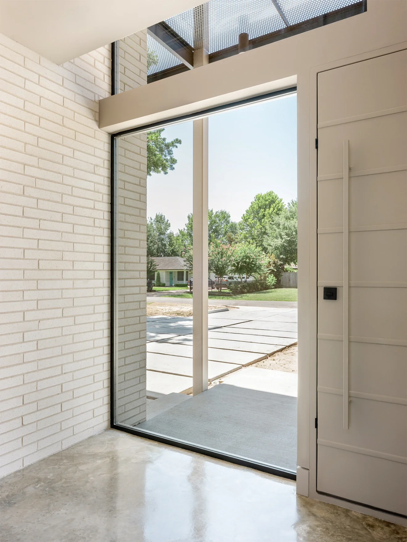 View from inside a modern house looking outside through a large glass door, showing trees, a street, and neighboring houses.