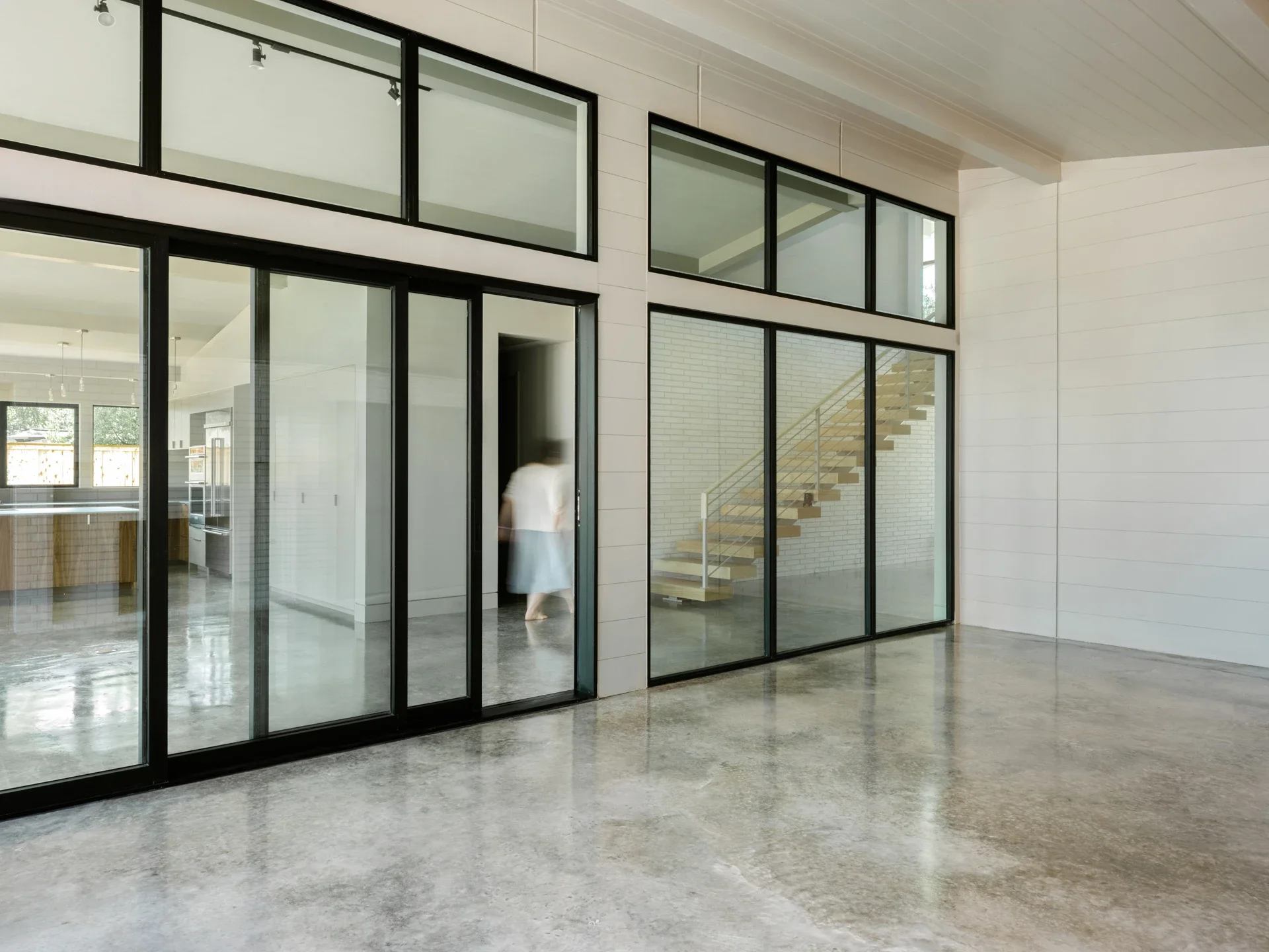 Modern interior space featuring large glass sliding doors and windows, concrete floor, white walls, and a staircase with wooden steps and a metal railing.