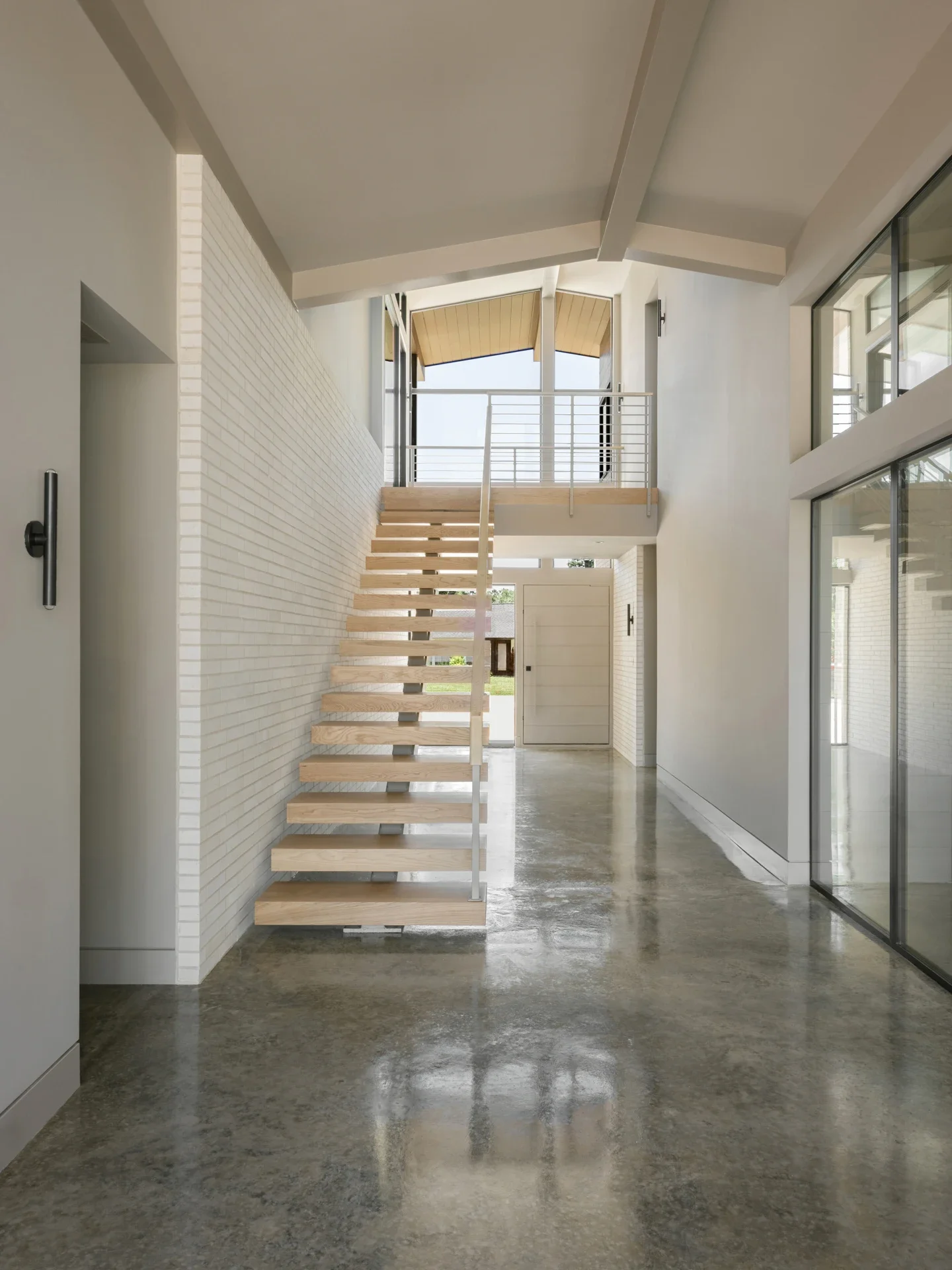 Interior view of a modern home with white walls, concrete flooring, a floating wooden staircase, and large windows.