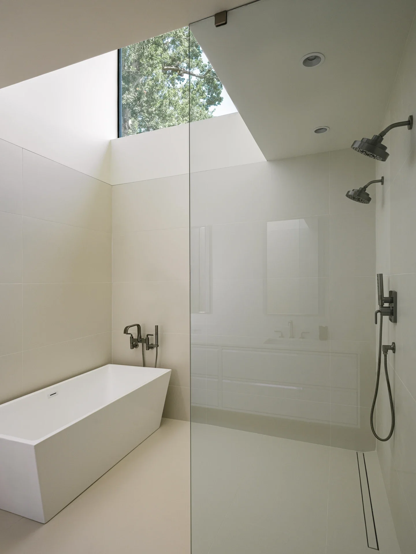 Modern bathroom with a freestanding bathtub on the left, a glass partition on the right, two ceiling shower heads, and a high window showing trees outside.