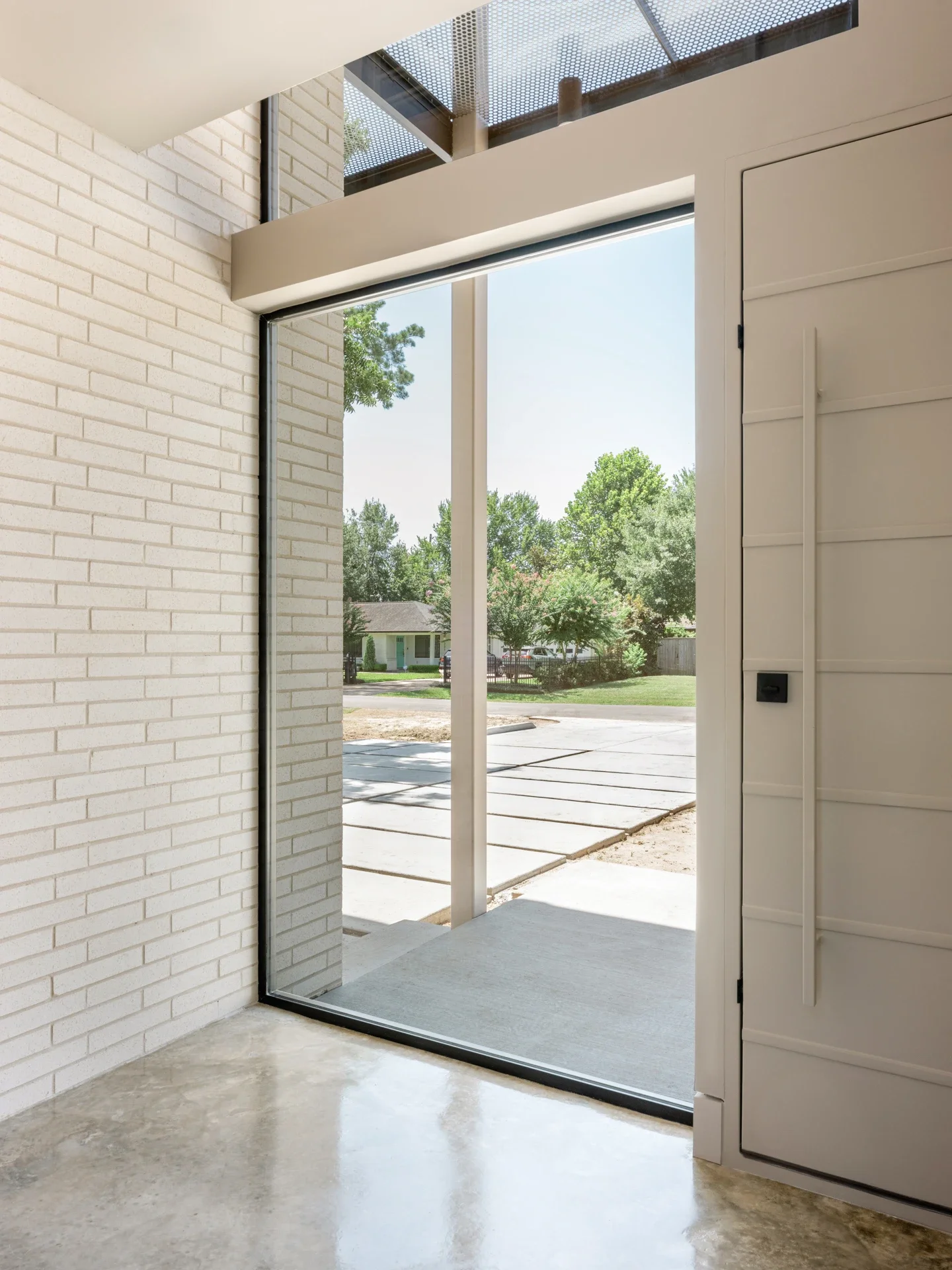 View of a modern house entryway with a large glass door and a concrete floor, overlooking a suburban street with trees and houses.