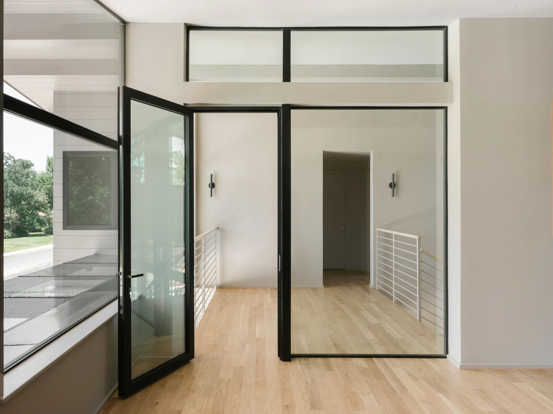 Interior view of a modern home with wooden flooring, large glass windows, and a glass door leading outside. There are white painted walls, black metal framing, a railing, and minimal decor.