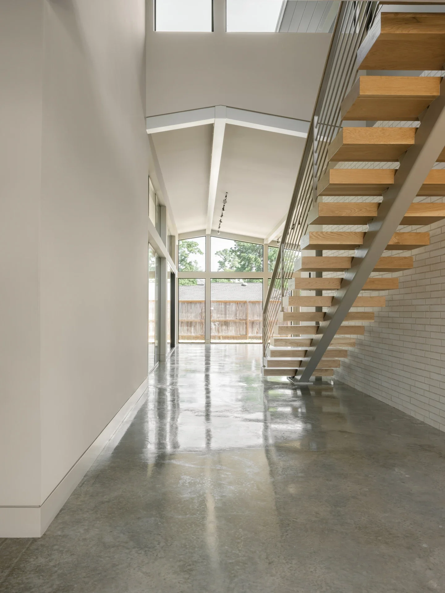 Modern interior of a house featuring a polished concrete floor, large windows allowing natural light, white walls, and a floating wooden staircase with metal railings leading to an upper level.