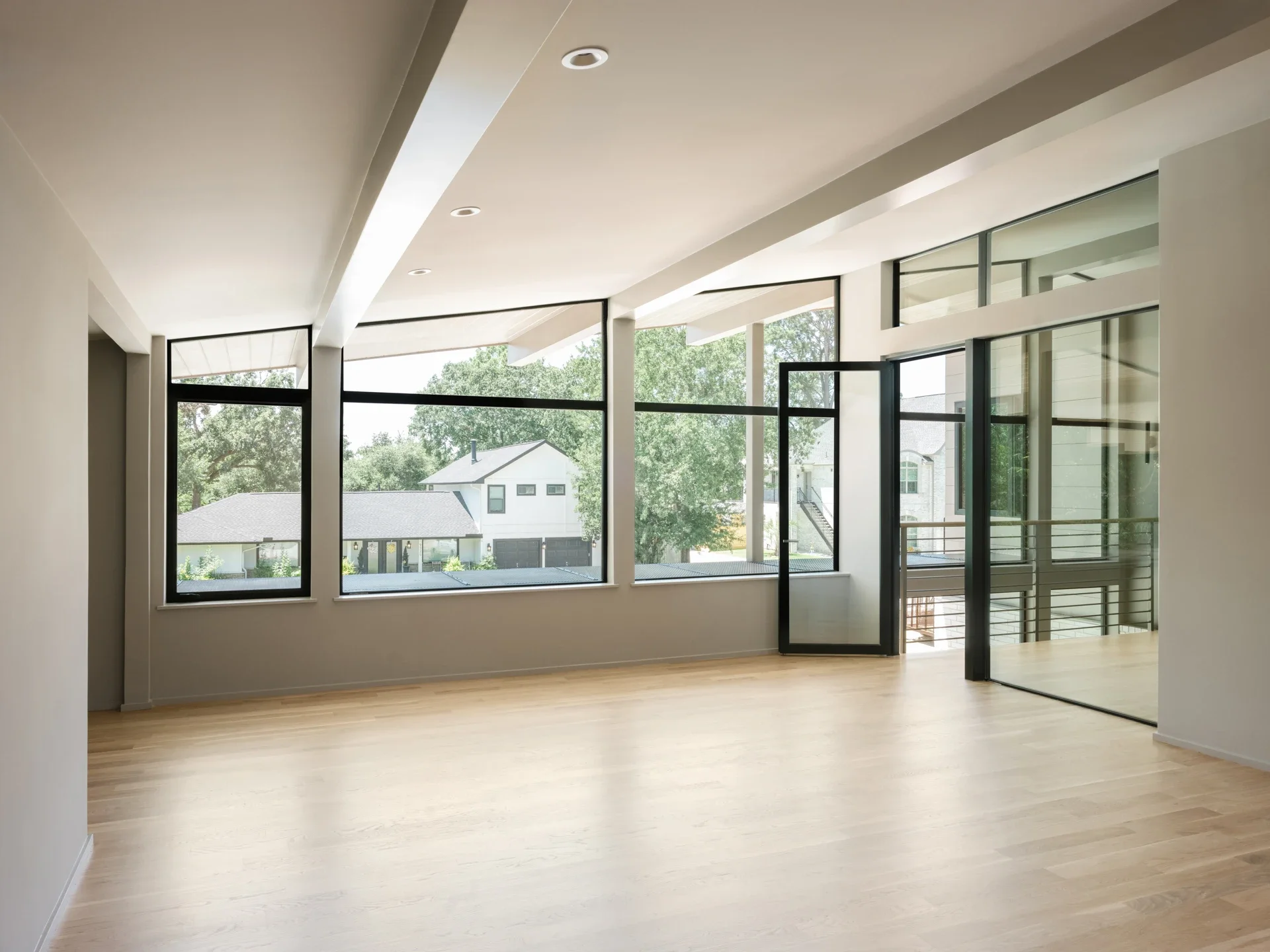 Empty room with large windows and sliding glass door, hardwood floors, and white walls, showing a view of neighboring houses and trees outside.