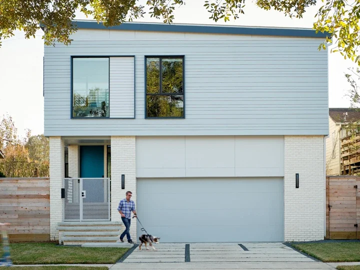 A modern two-story house with a light blue upper level, white brick lower level, and a large garage door. A man walks a dog on the driveway.