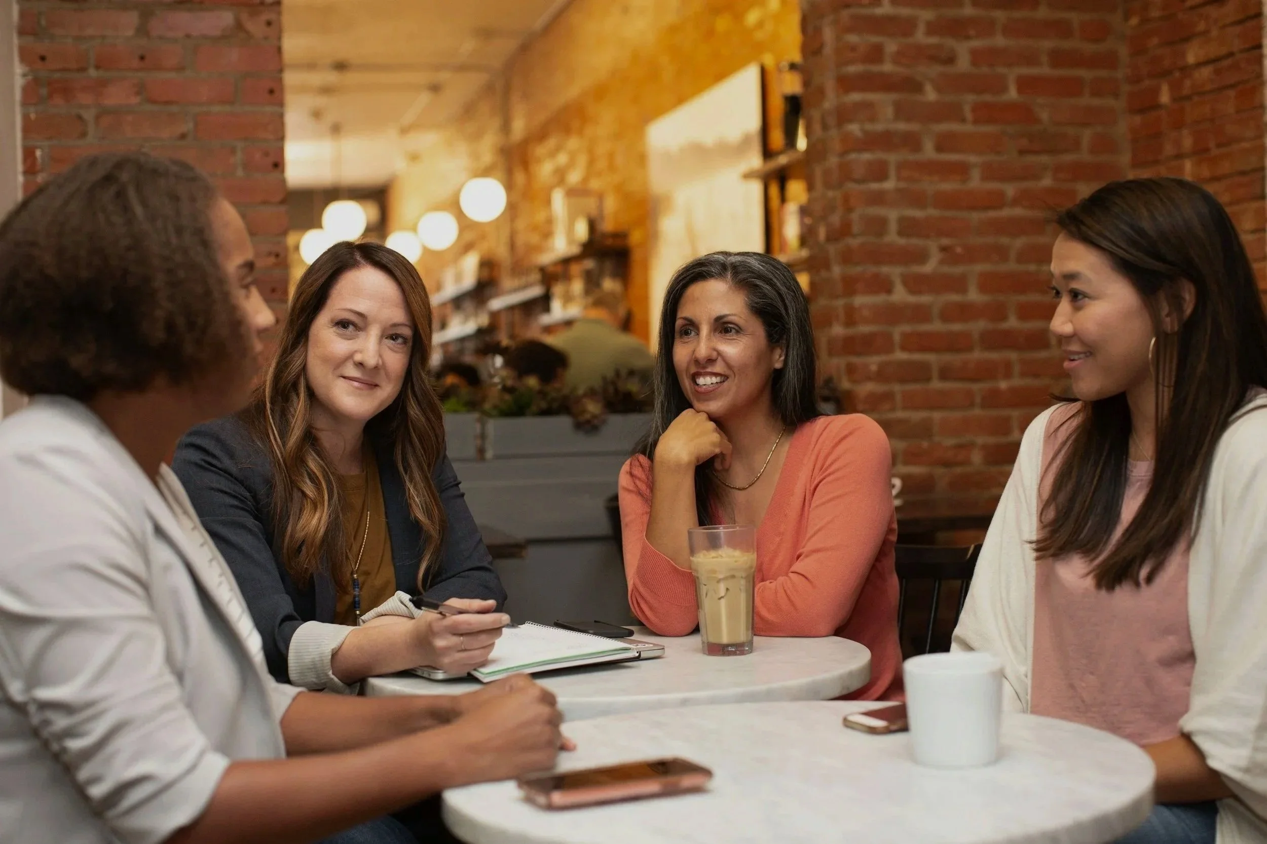 Four women sitting at a small round table in a cafe, engaged in conversation. One woman is holding a pen and notebook, another has a tall glass of iced coffee, and there are smartphones and a mug on the table. The background features a brick wall and warm lighting.
