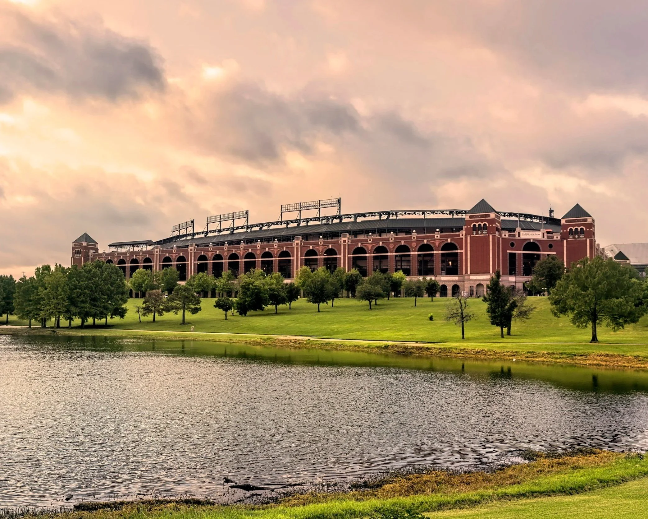 A large, brick stadium situated on a grassy hill with trees, next to a body of water under a cloudy sky.