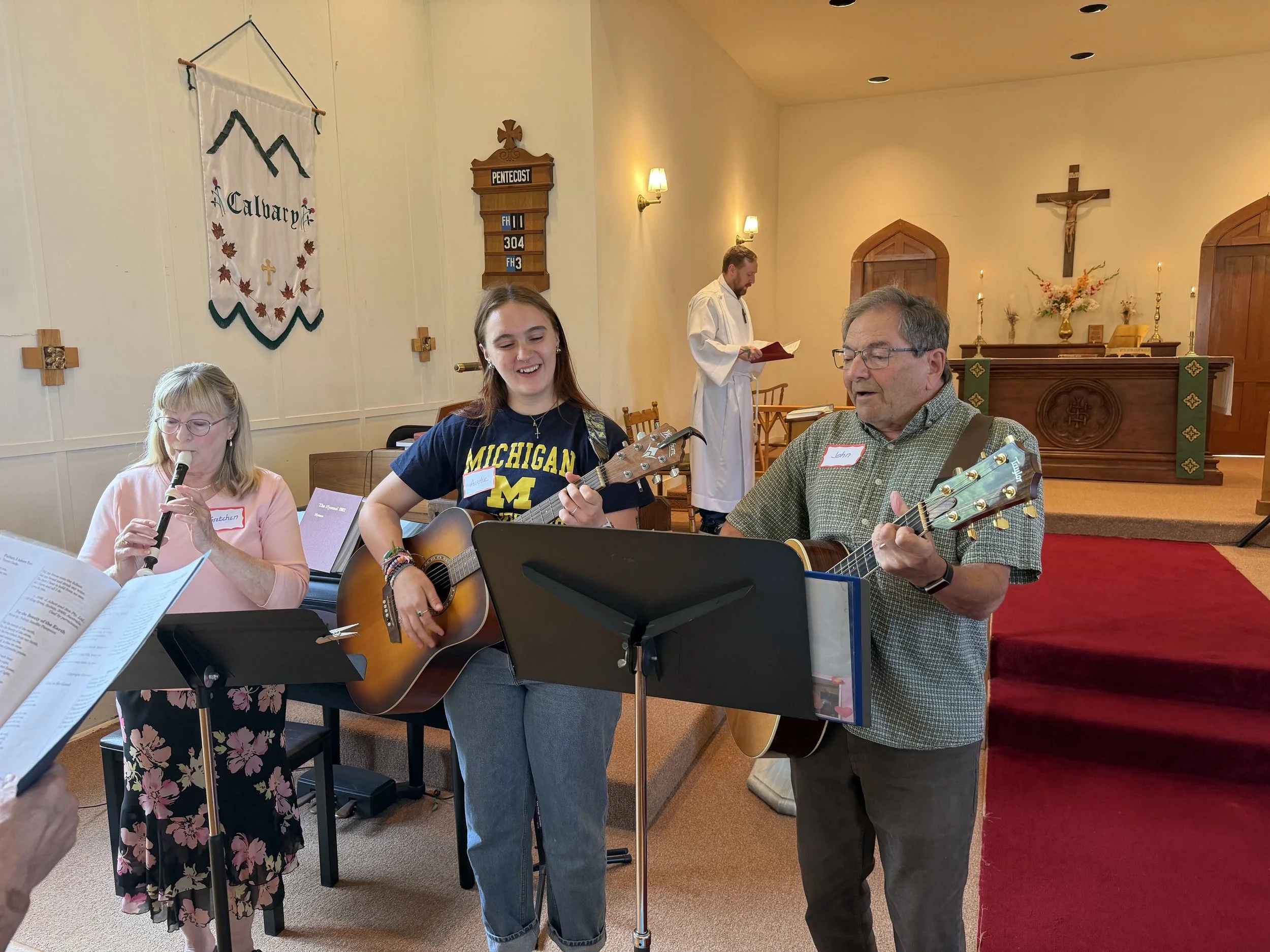 A group of people participating in a musical event inside a church, with a woman playing a recorder, a young woman playing a guitar, and an older man also playing a guitar. They are standing in front of the altar, which is decorated with candles, flowers, and a crucifix on the wall.