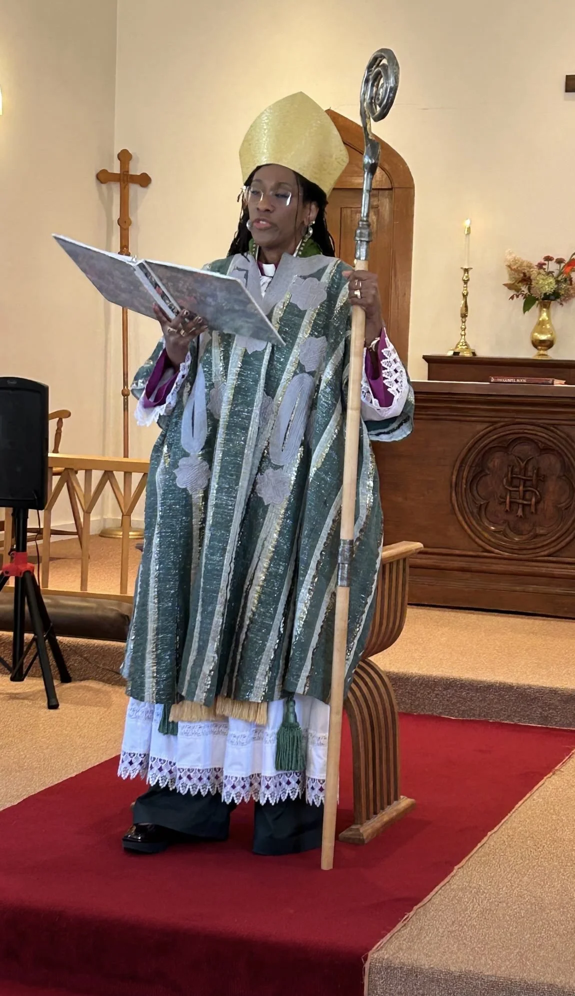 Woman Bishop standing in front of church alter holding staff reading to congregation. Wearing golden headdress and green and white striped vestment.