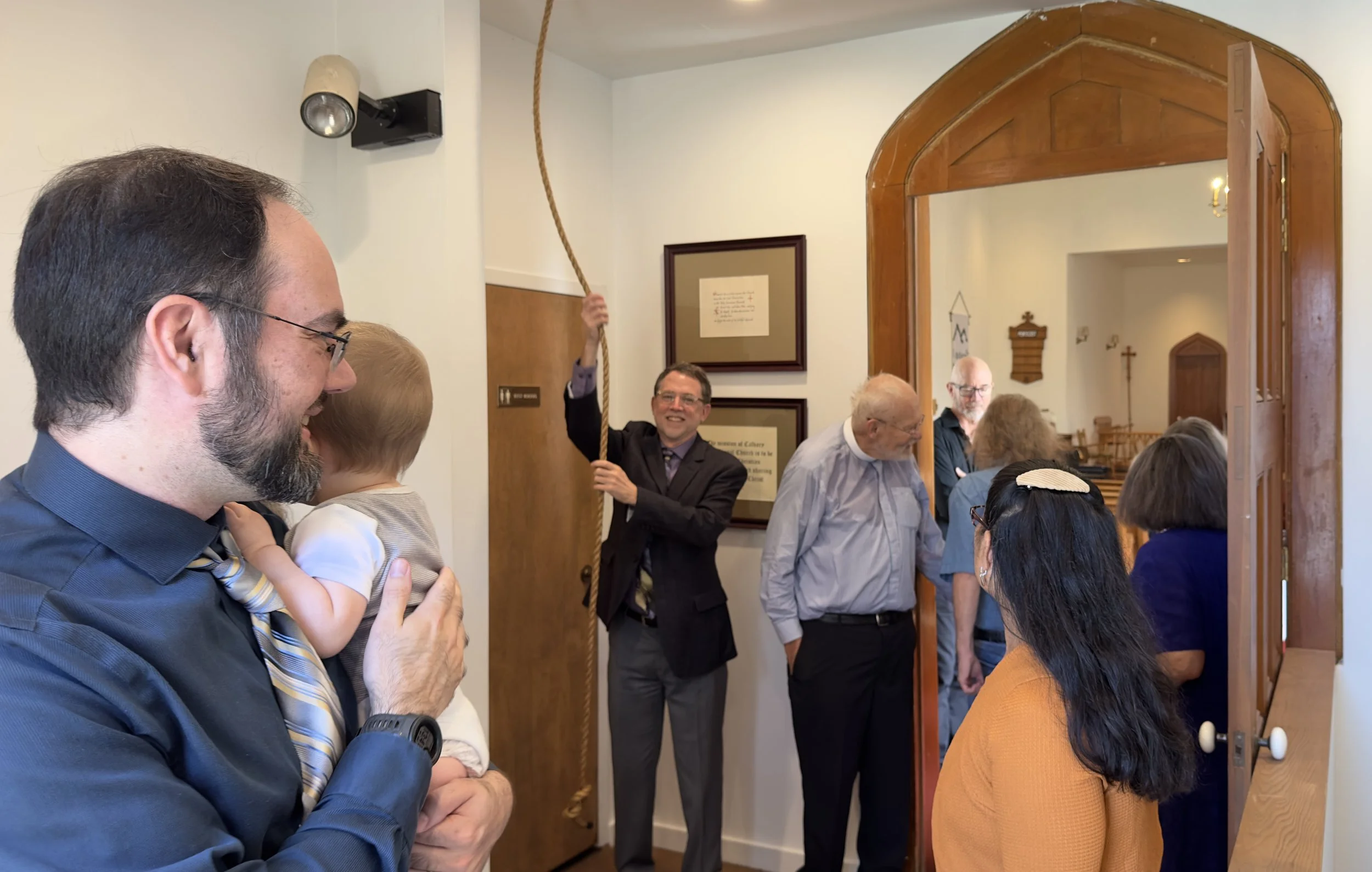 Man smiling in jacket and tie pulling rope to ring the church bell. A man with baby looks on.