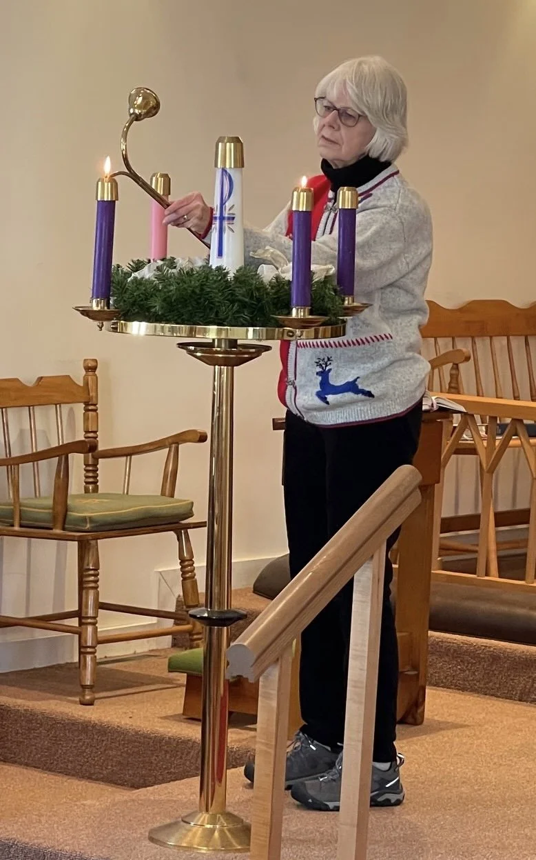 Female parishioner wearing a gray and red Christmas sweater with reindeer design lighting an advent calendar with long brass candle lighter. There are 5 candles on the brass candle holder decorated with Christmas greenery.