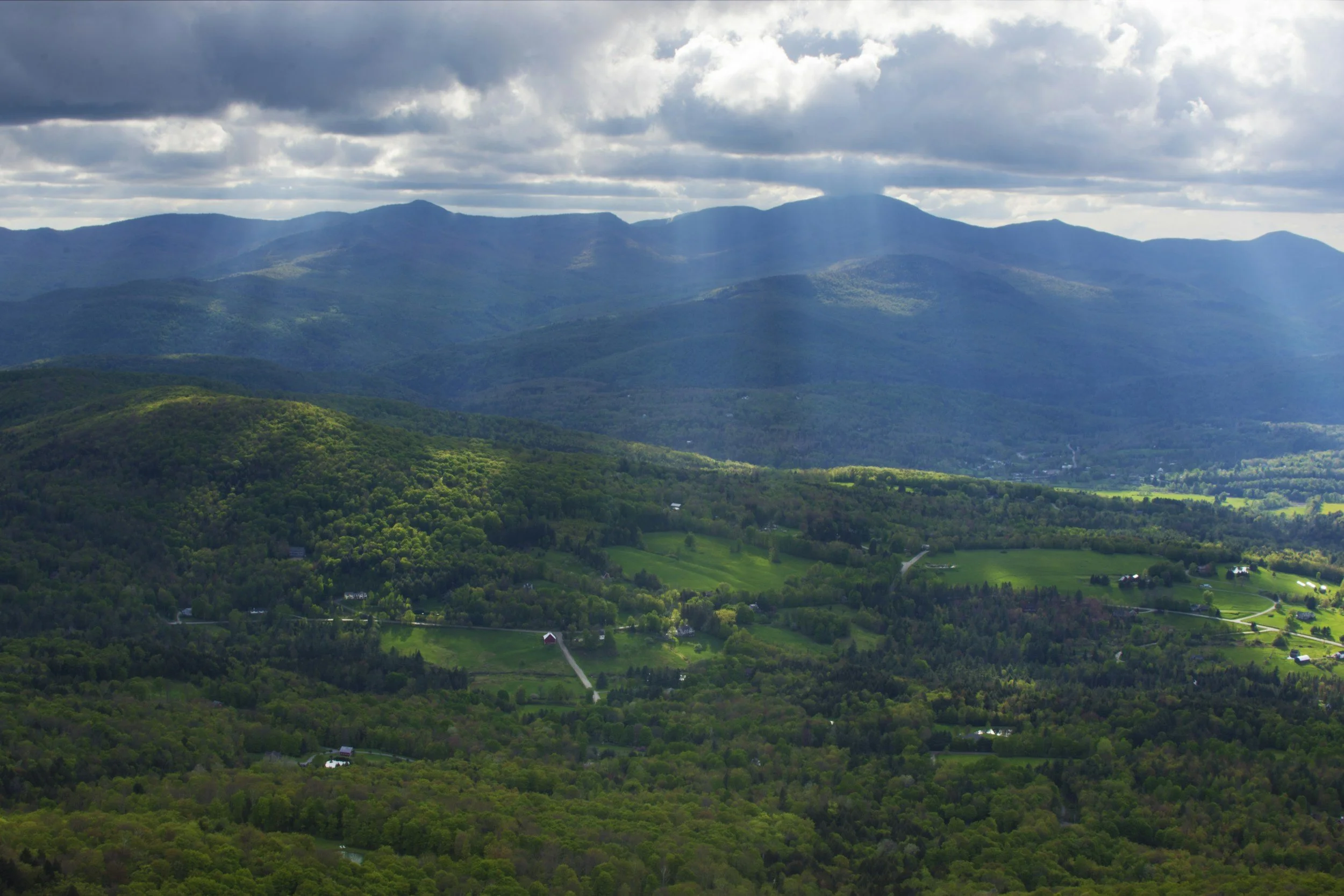 Vermont summer landscape with wooded forests, green fields, the Green Mountains, and a dramatic sky with dark clouds and rays of sun shining pouring down onto the land.