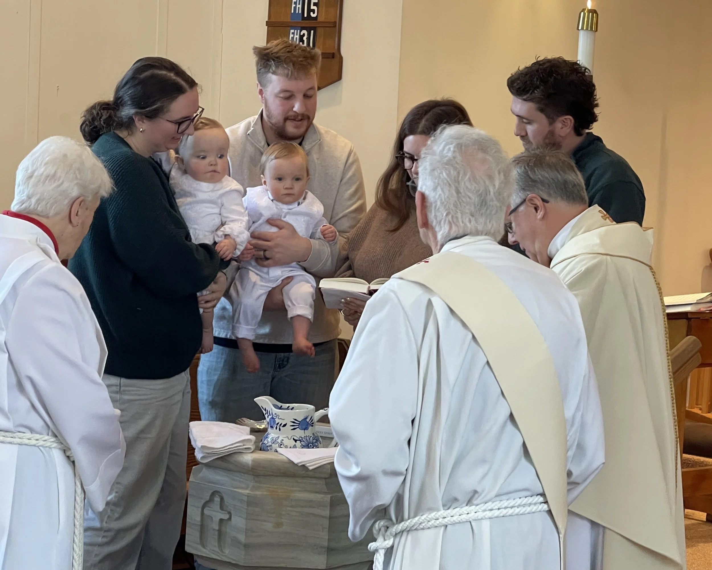 A baptism ceremony with twin babies, Mom, dad, godparents, and three clergy members surrounding the baptismal font.