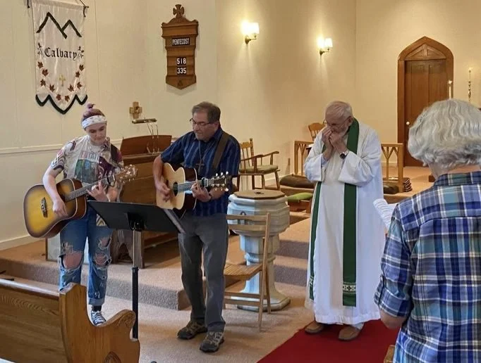 Three musicians: A man and woman playing guitars and an older priest in white vestment and green stole playing harmonica.