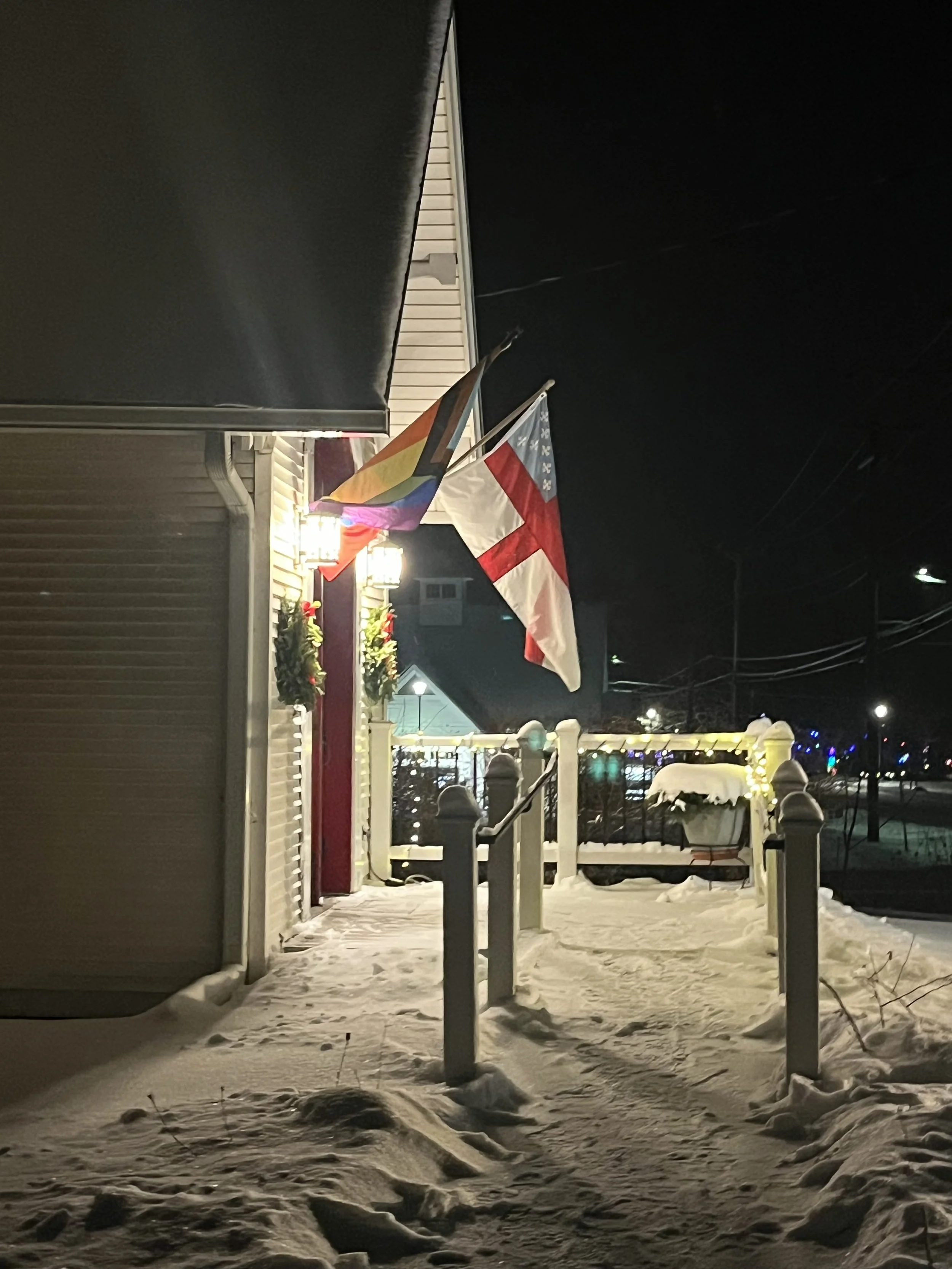Outside of Calvary Church at night in the snow with lights, Episcopal flag, pride flag, green wreaths, and red door.