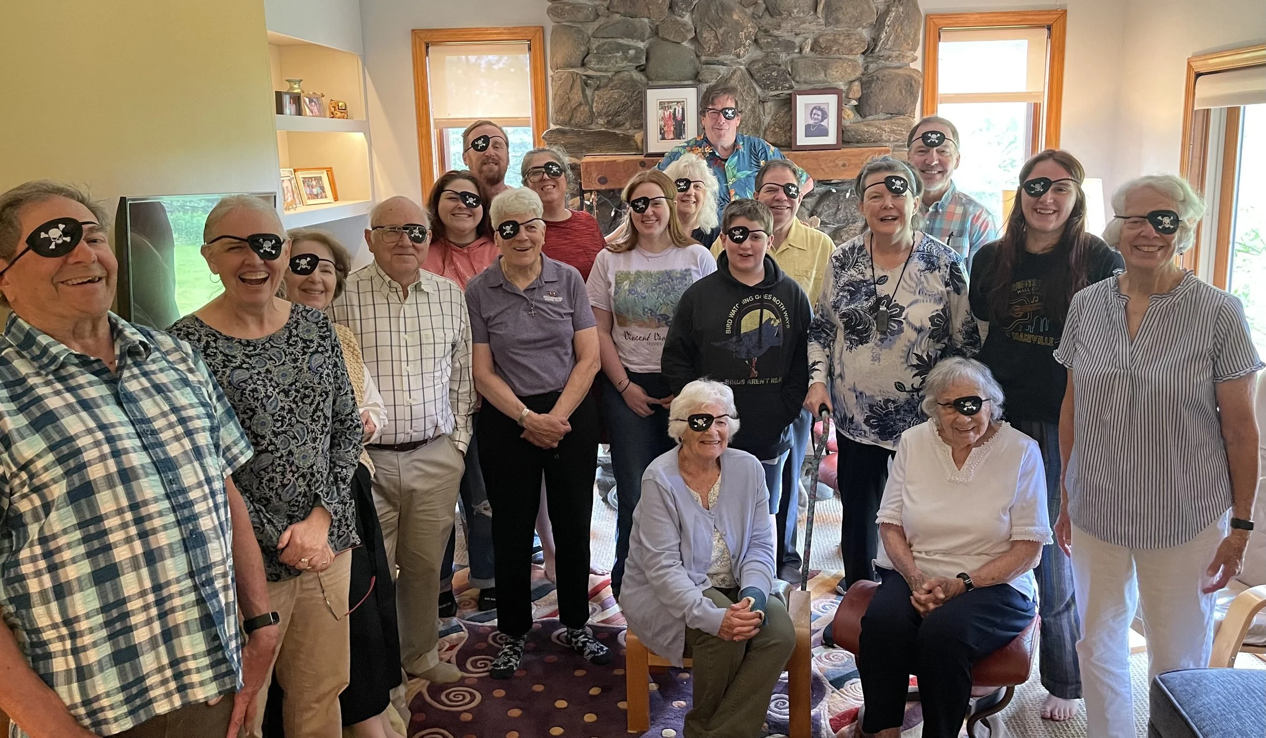 A group of people wearing pirate eye patches with skull and crossbones in a living room with multiple windows, a stone fireplace, and framed photos on the wall.