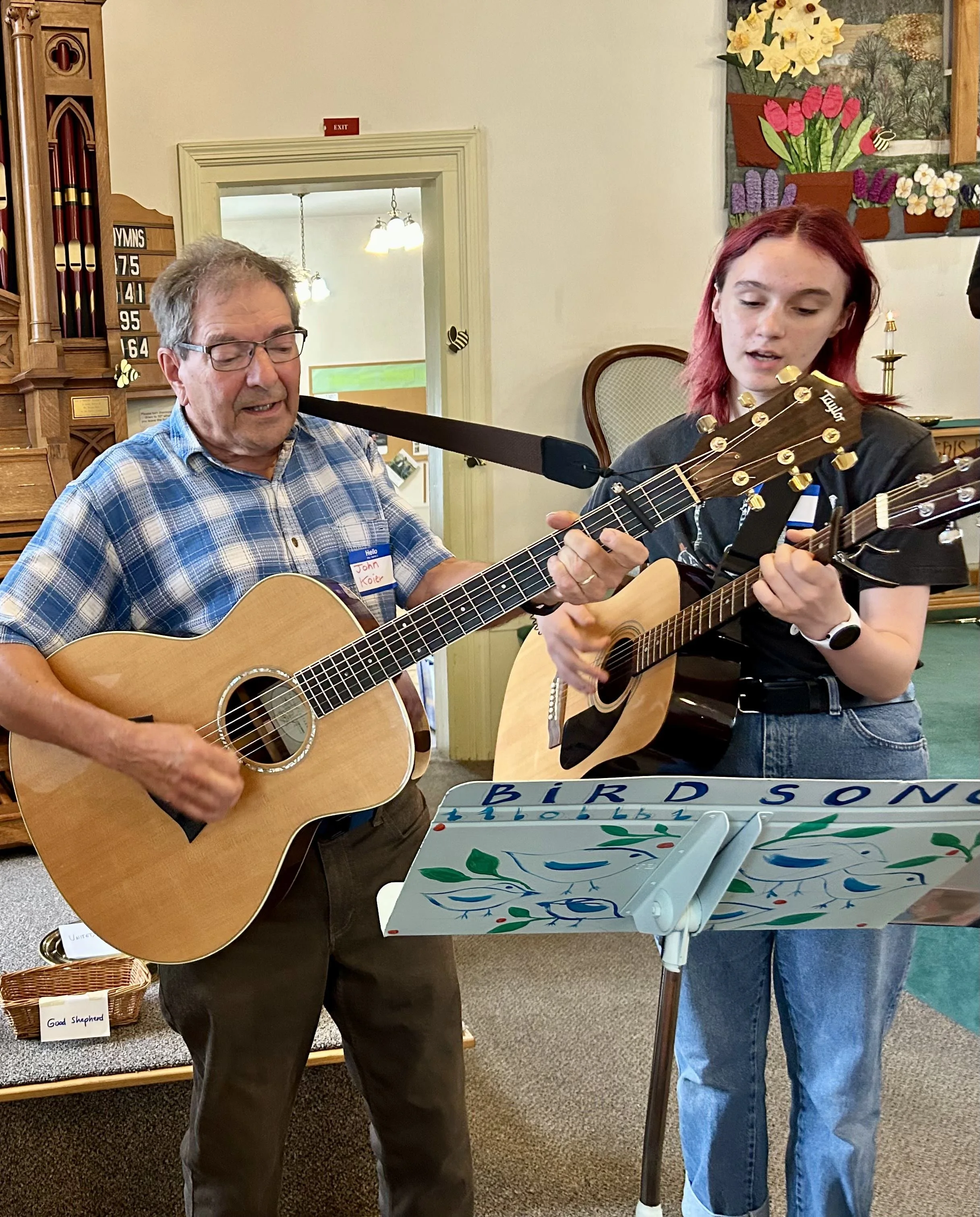A young woman and older man playing guitars. Music stand has painted vines and says BIRD SONG. There is a sewn bright floral tapestry behind them.