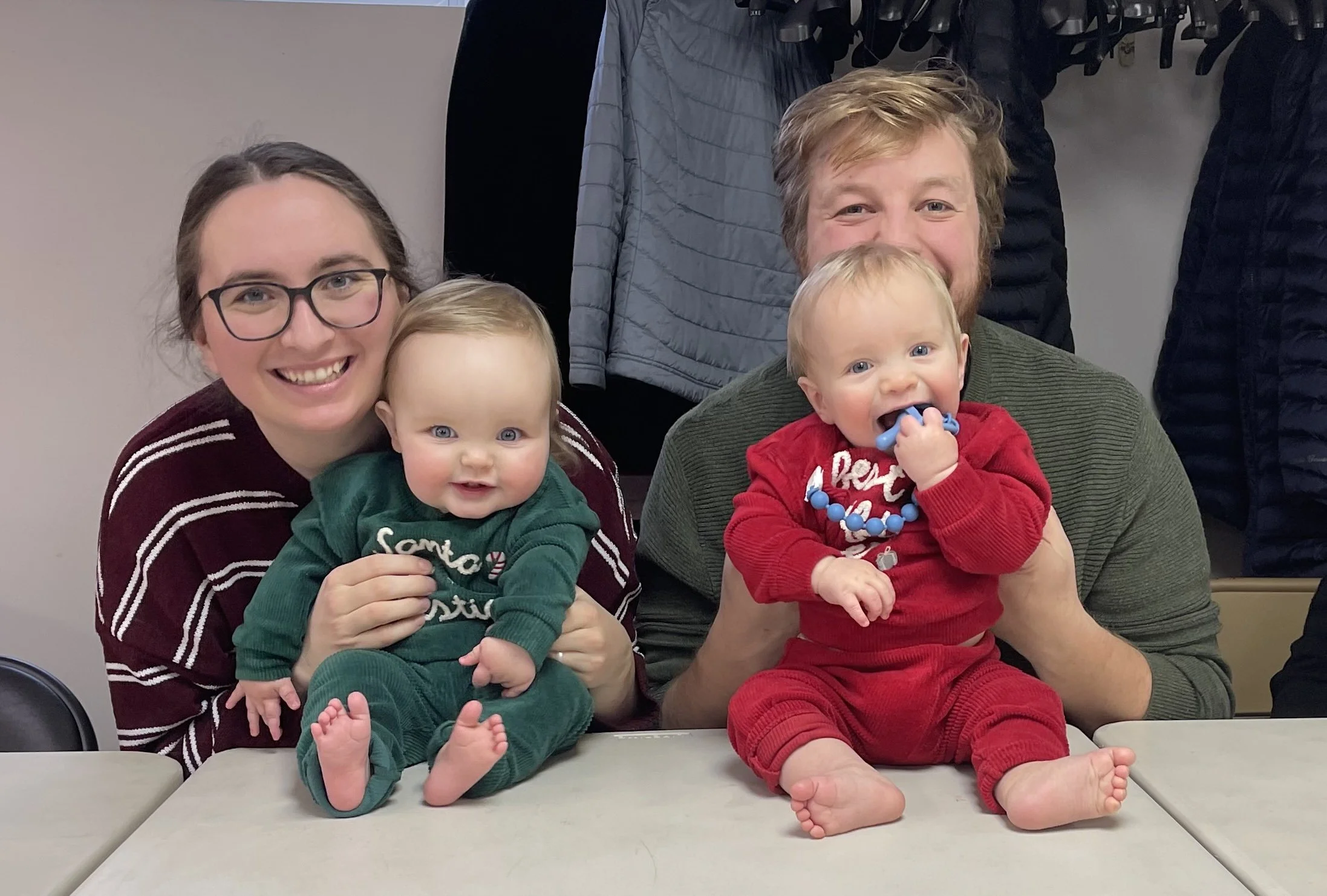 Smiling mother and father holding up twin babies who are sitting on a table. The twins are wearing Christmas outfits, one in red and the other green.