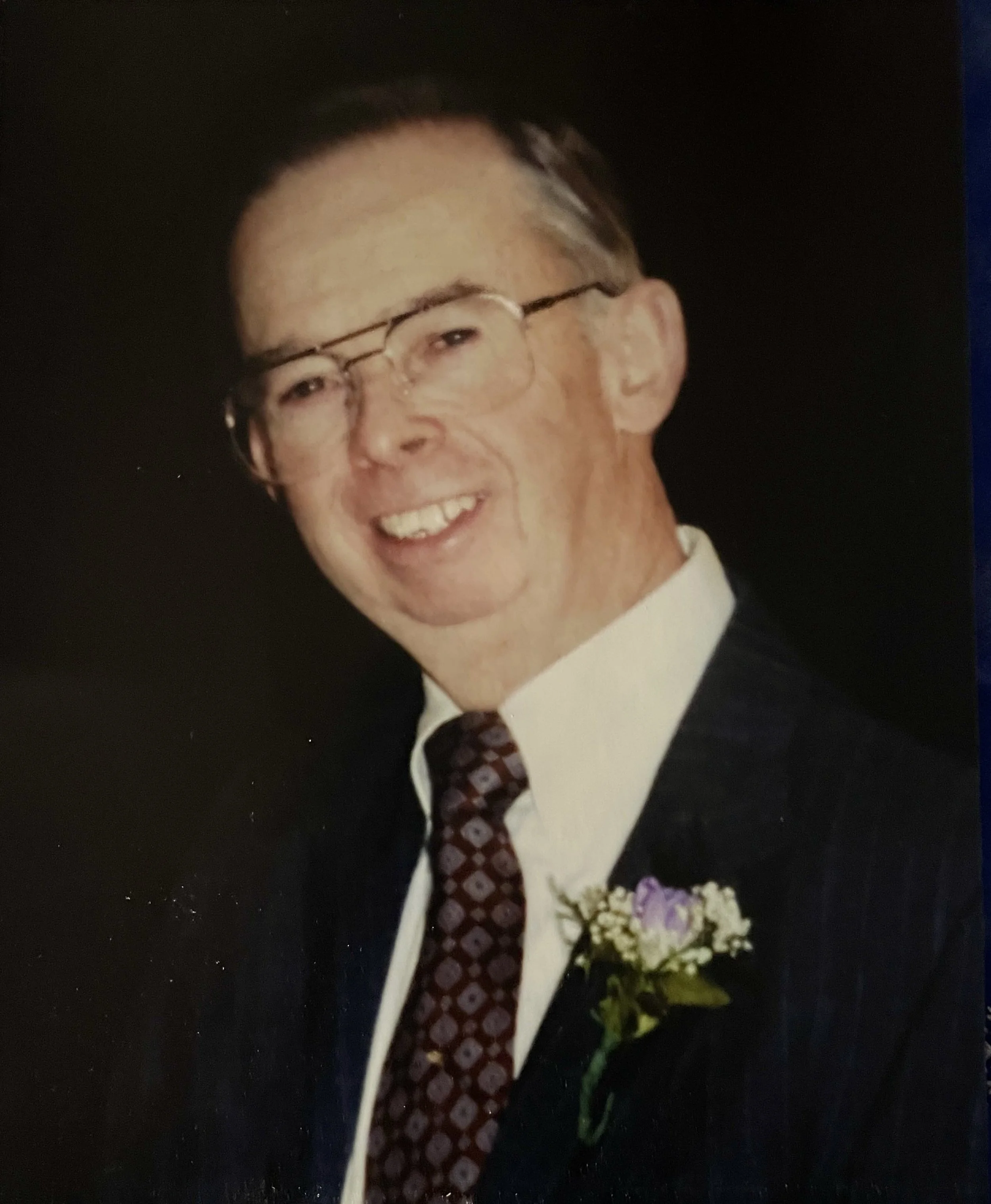 Smiling man wearing glasses, white shirt, rtie, and black jacket with boutonnìere.