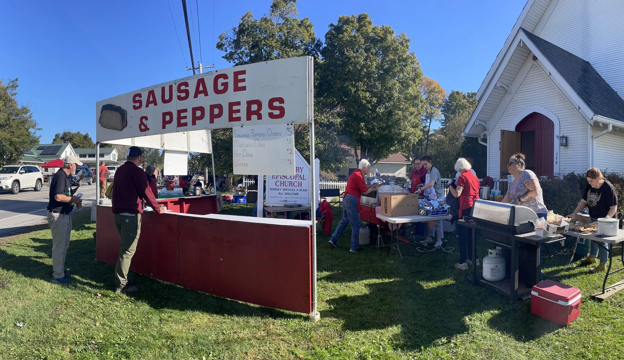 Calvary church with wooden stand out front and sign reading “Sausage & Peppers.” The stand and sign are red and white.