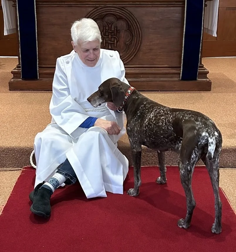 Woman and dog sitting on floor in front of alter. Woman is wearing a white vestment and dog is brown with white spots.