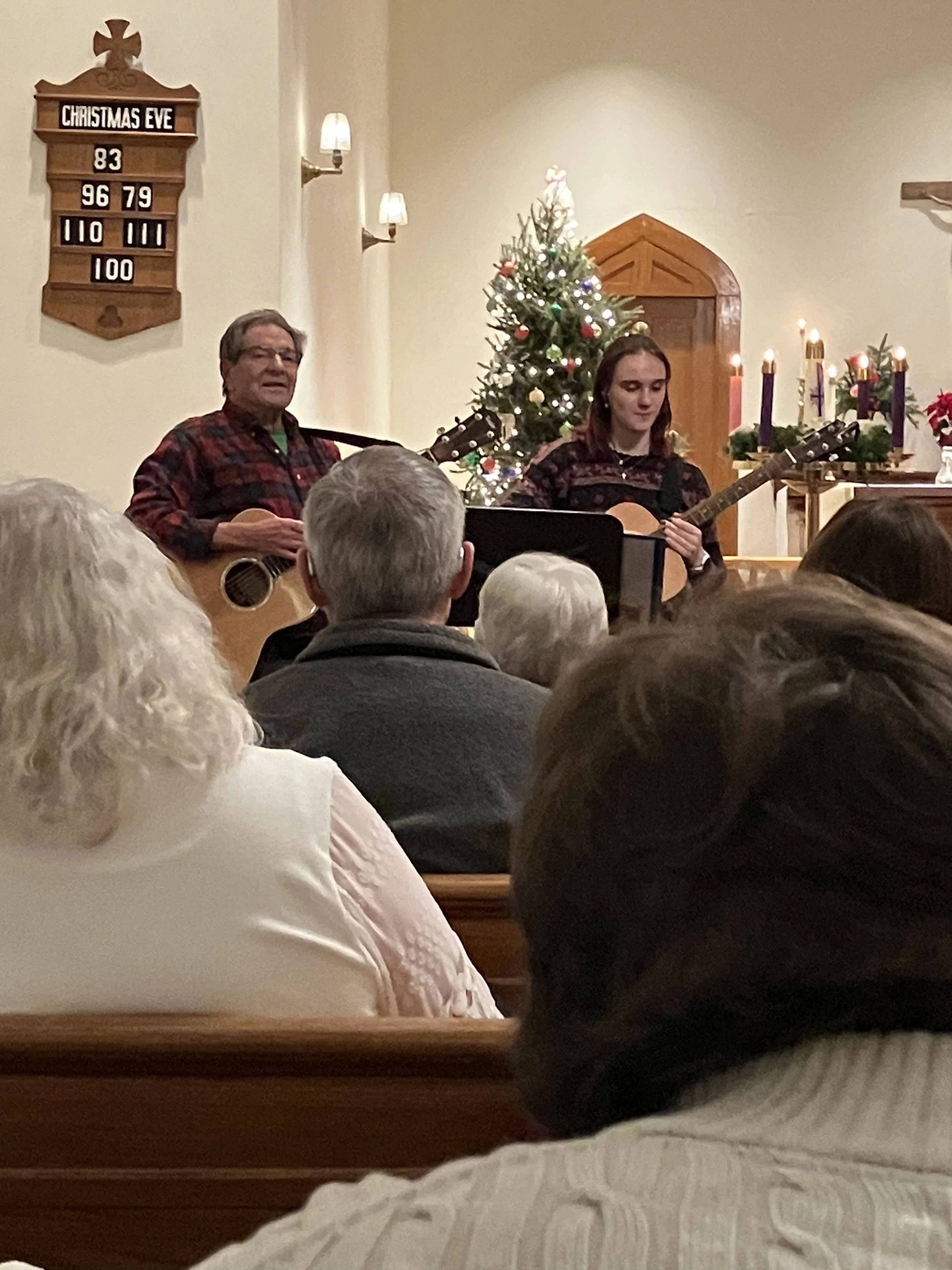 Two people playing guitars in a church during a Christmas Eve service, with a decorated Christmas tree, lit candles, and an Advent wreath with purple and pink candles in the background, and a hymn board on the wall.