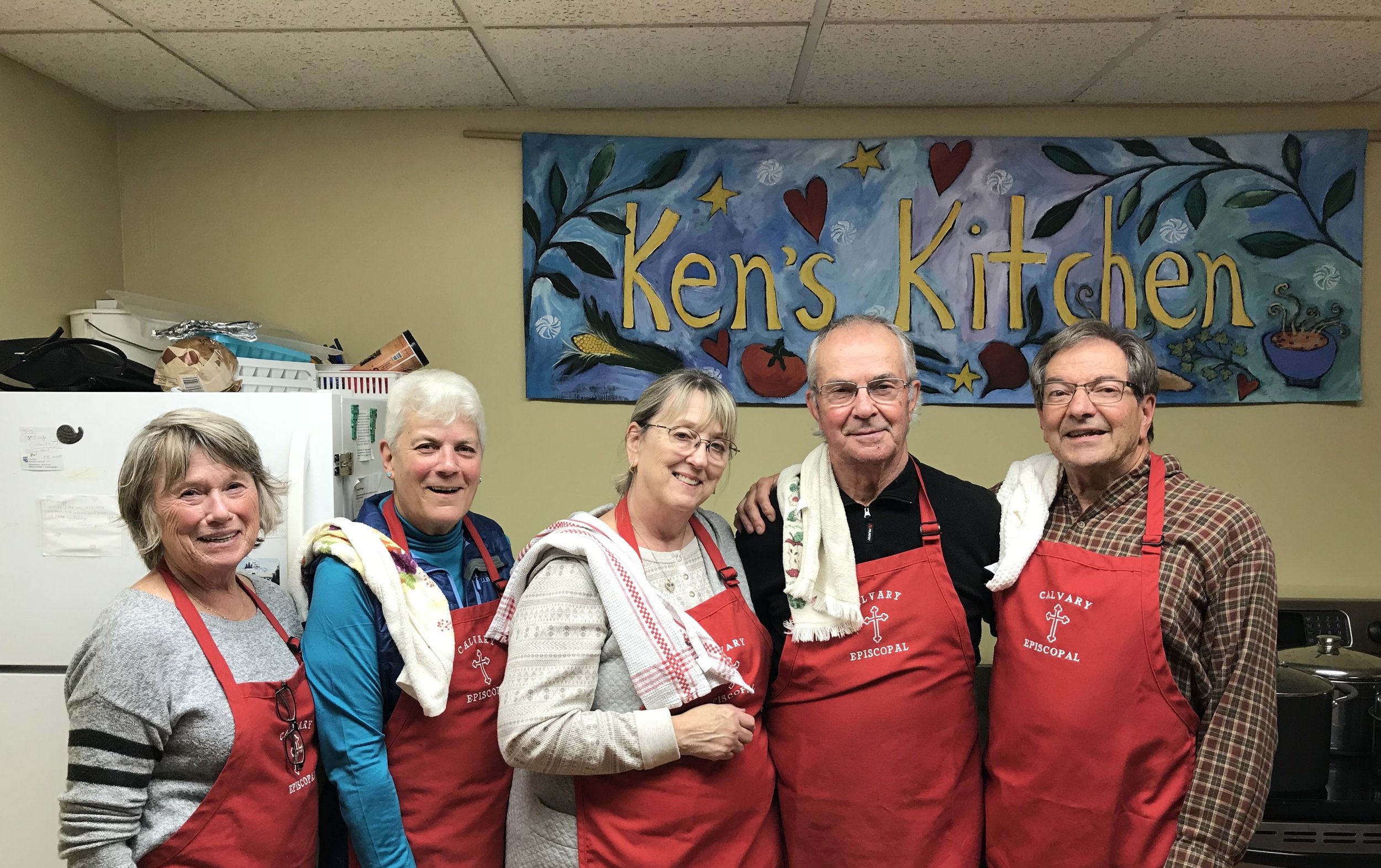 Five soup supper volunteers wearing red aprons with dish towels on their shoulders.