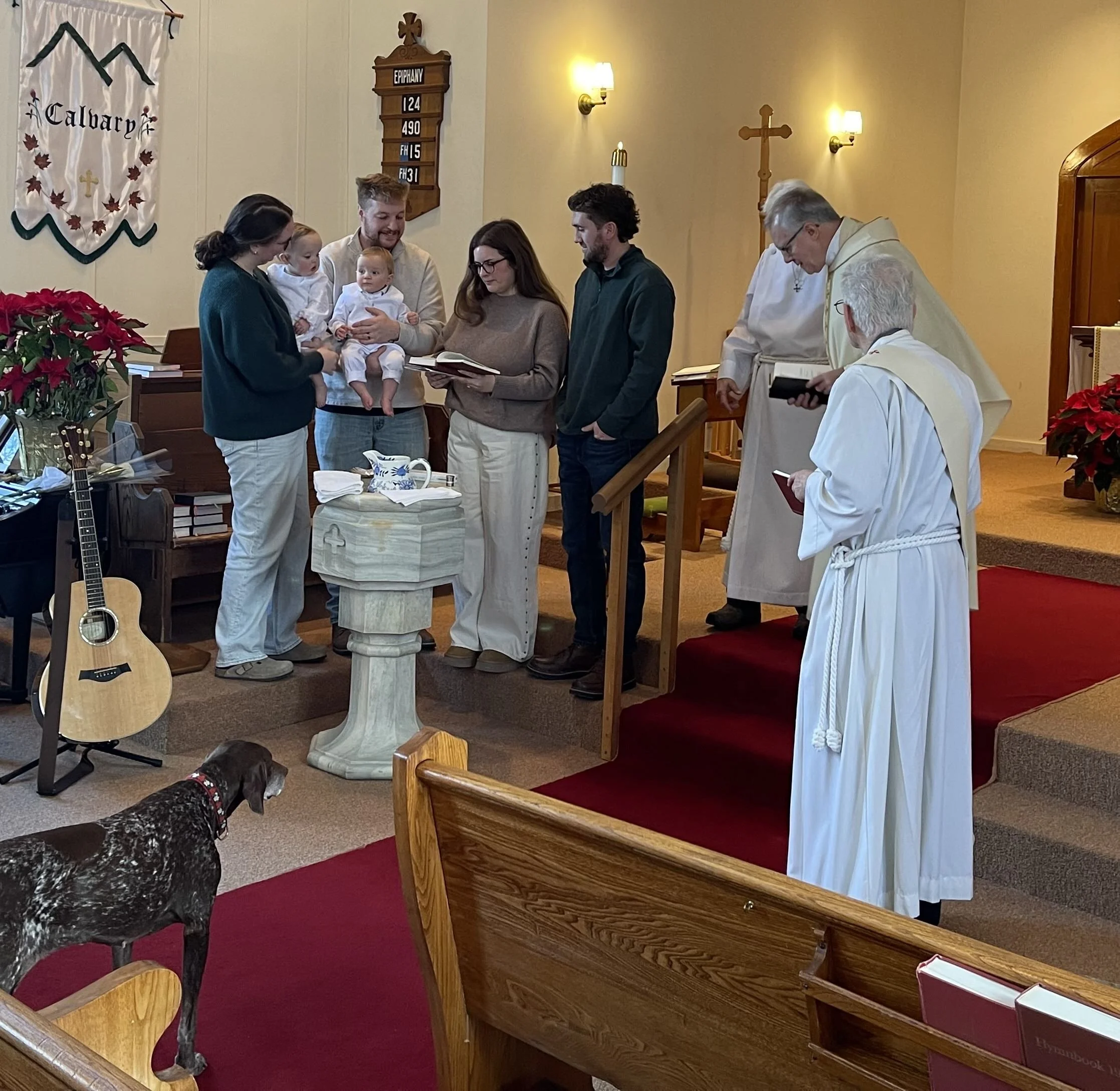 A baptism ceremony with twin babies, Mom, dad, godparents, and three clergy members surrounding the baptismal font with clergy standing to the side.