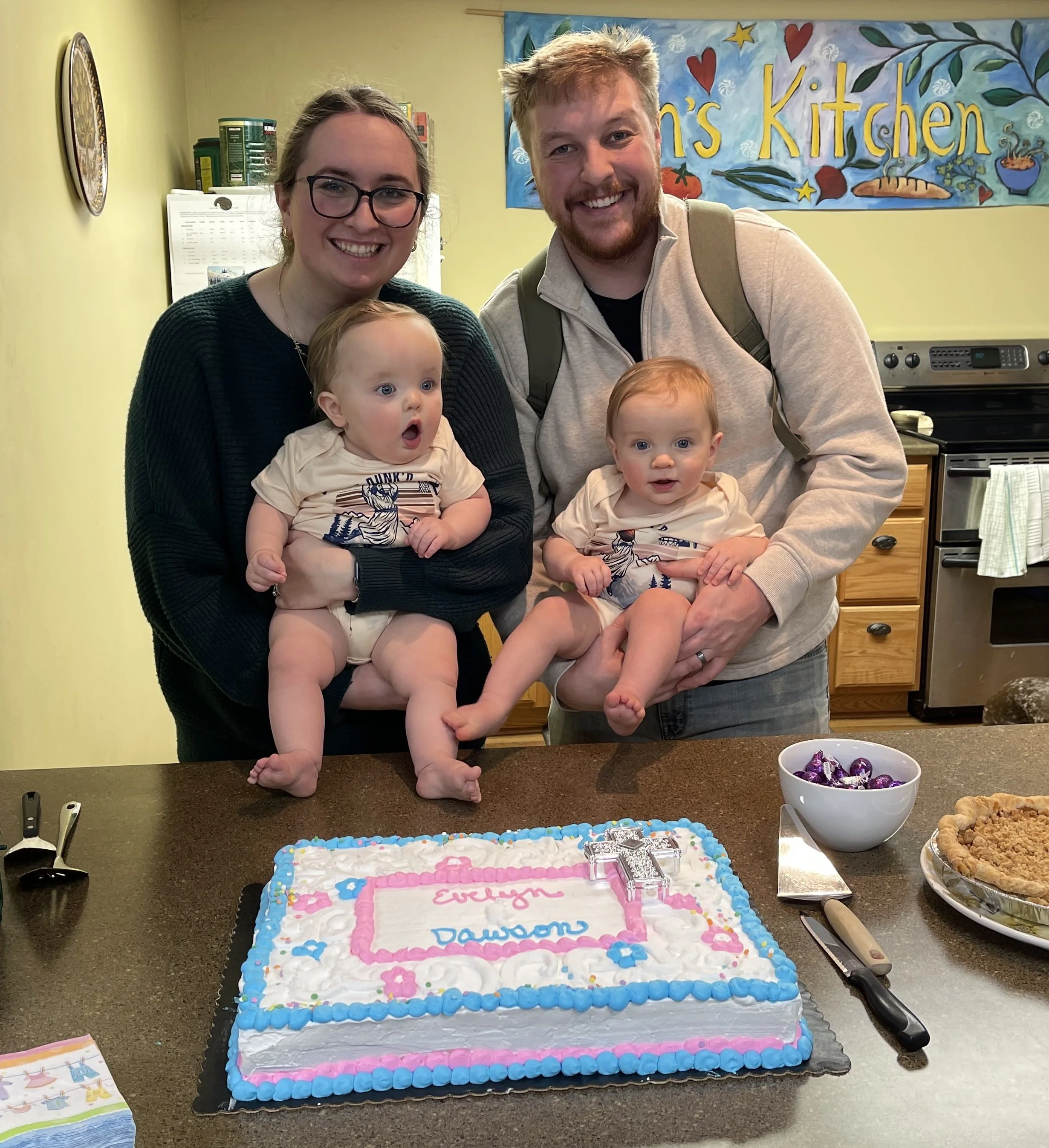 Mom and dad holding twin babies in front of a cake to celebrate their Baptism.