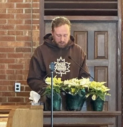 Young male parishioner wearing a brown hoody giving a reading behind the lectern at St. James Church in Essex. There are three pale green poinsettias on the wooden lectern.